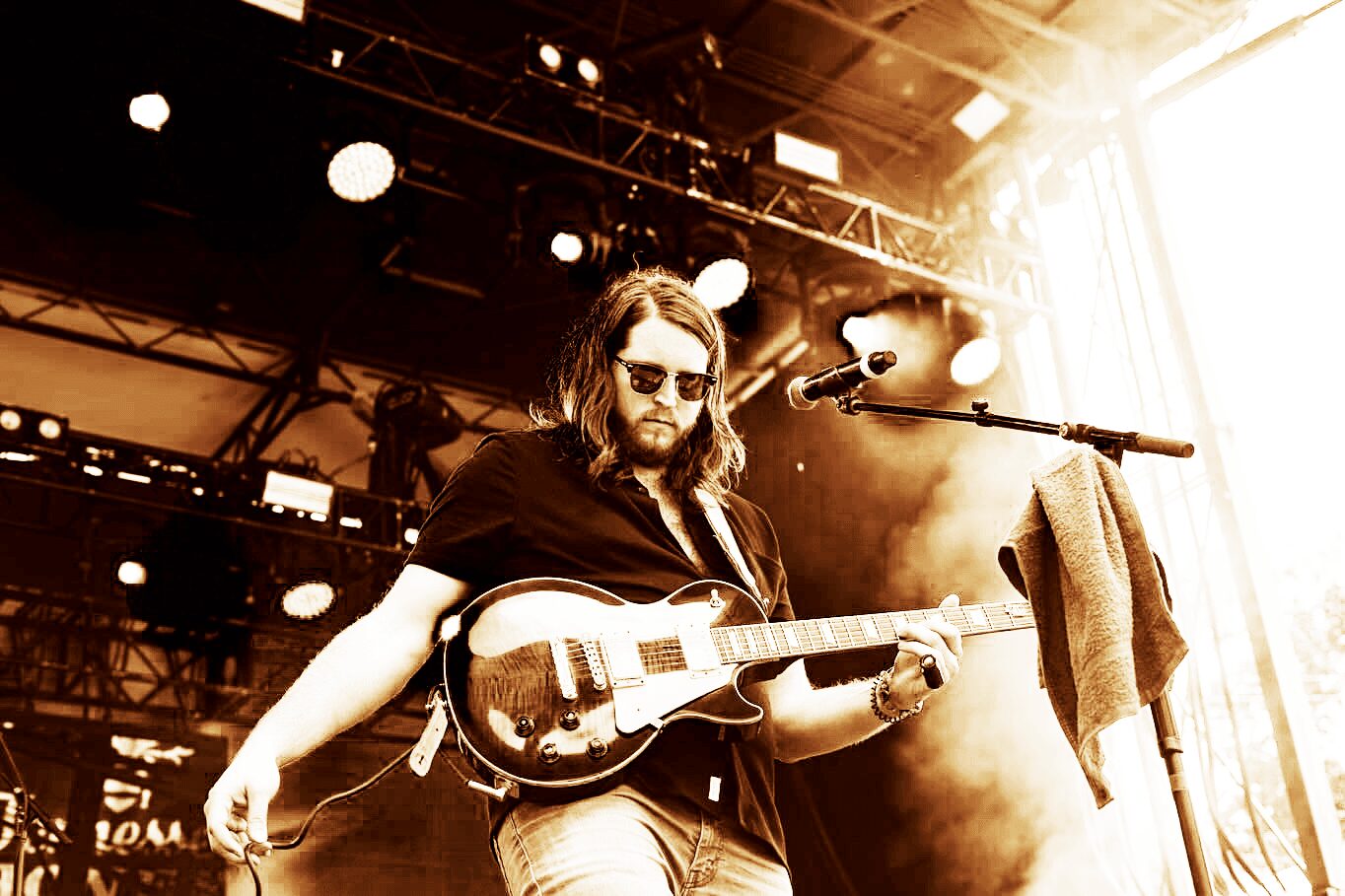 Musician with long hair and sunglasses playing an electric guitar on stage with bright lights and equipment behind him.