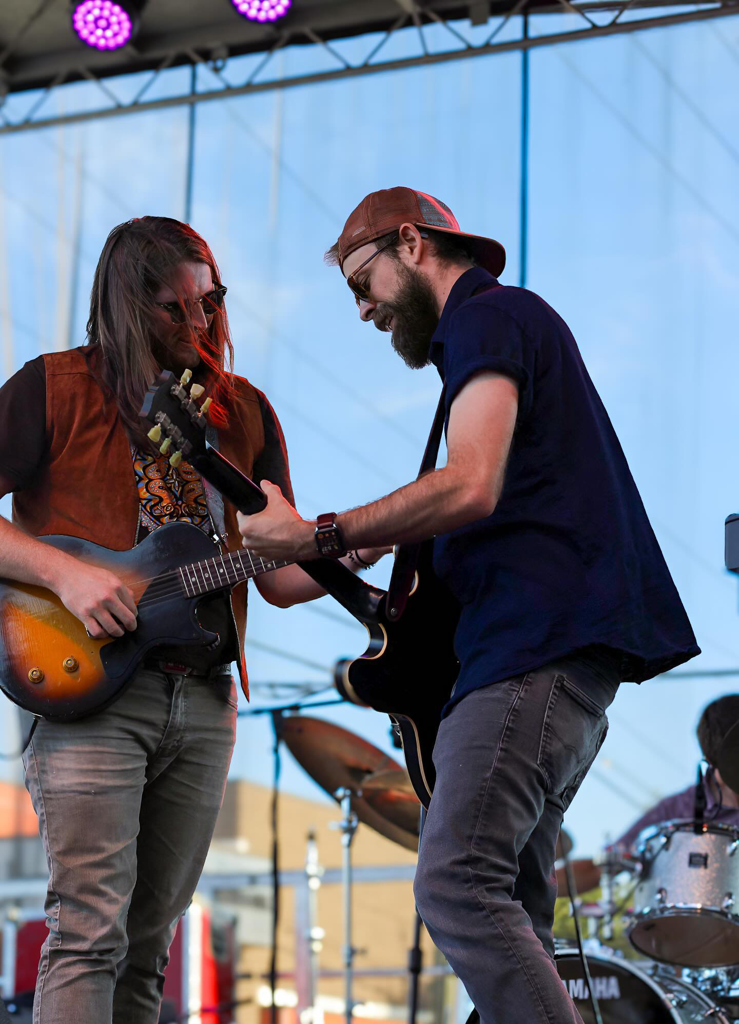Two musicians playing guitars on stage, one with long hair and the other with a cap, with a drum set in the background.