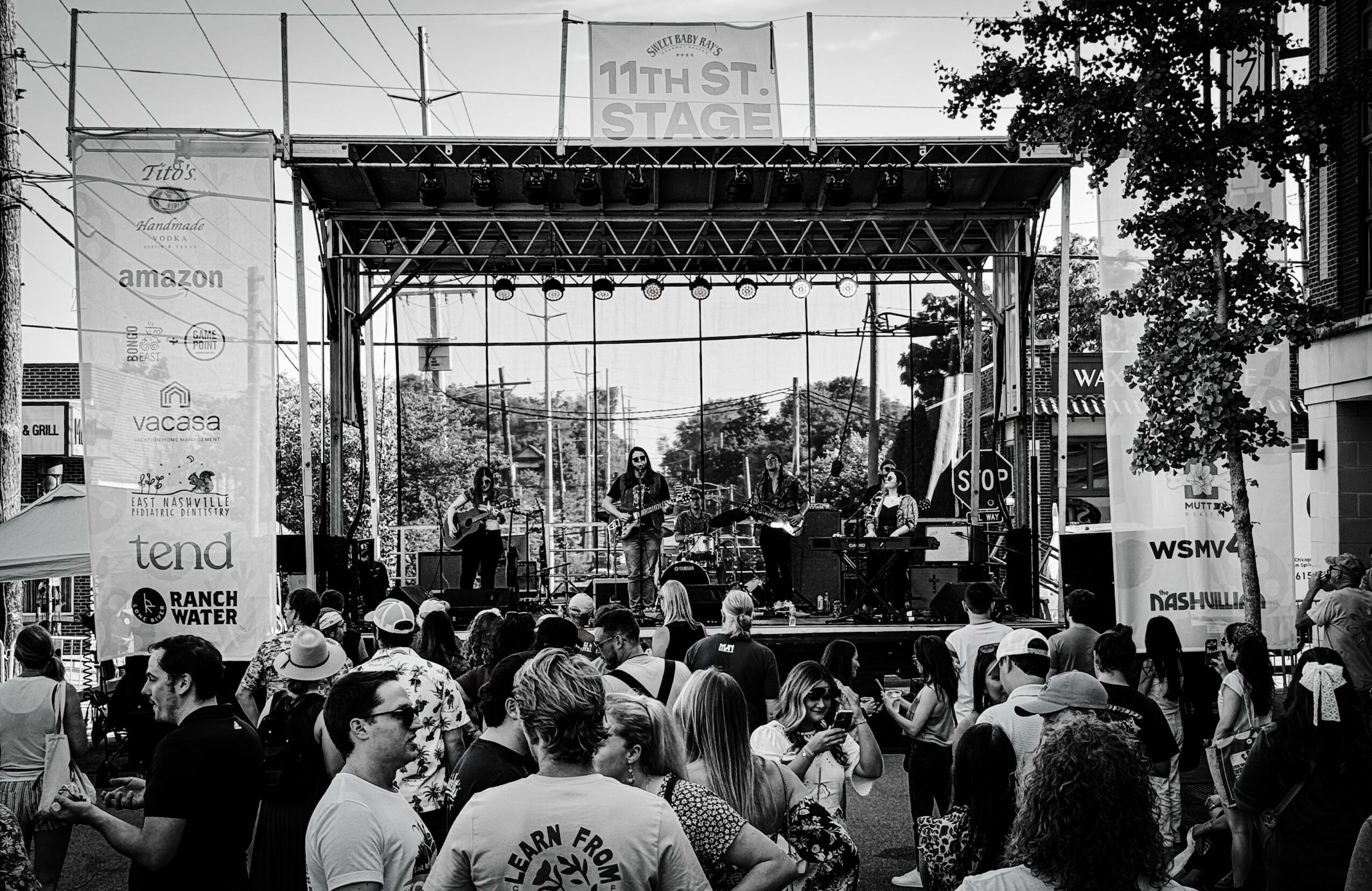 Crowd watching a live outdoor concert on a stage with musicians, banners, and trees in the background.
