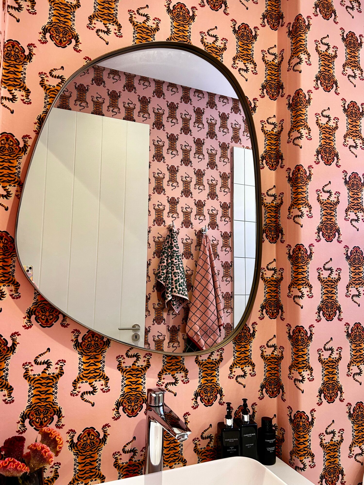 Oval mirror reflecting a wall with tiger-patterned wallpaper and a door, with a person and clothing visible in the reflection.