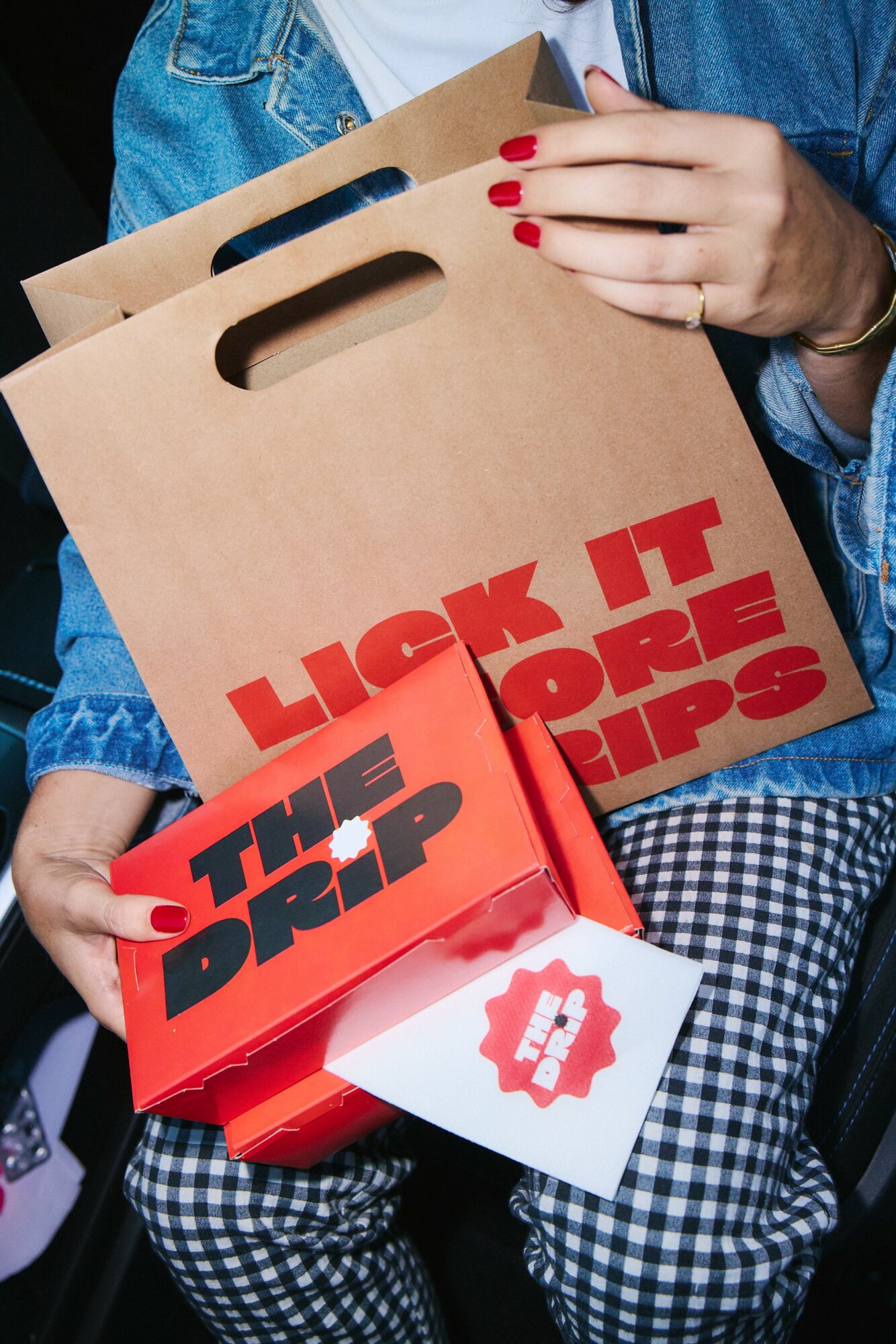 Person holding shopping bags with red text, one bag says 'LICK IT BEFORE YOU TRIP,' and the other says 'THE DROP.'