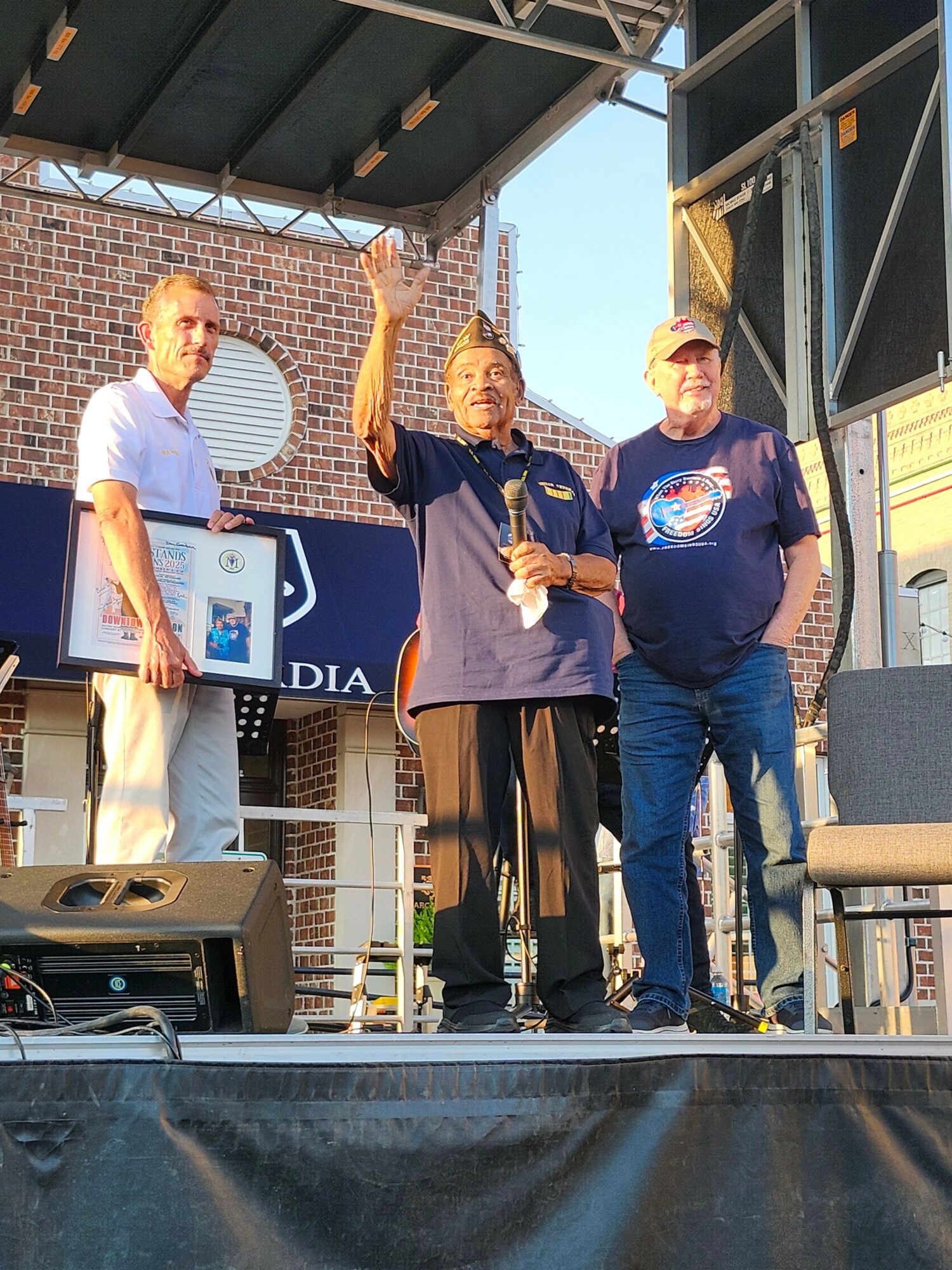 Three people standing outdoors, one holding a framed certificate, with a brick building and cityscape in the background.