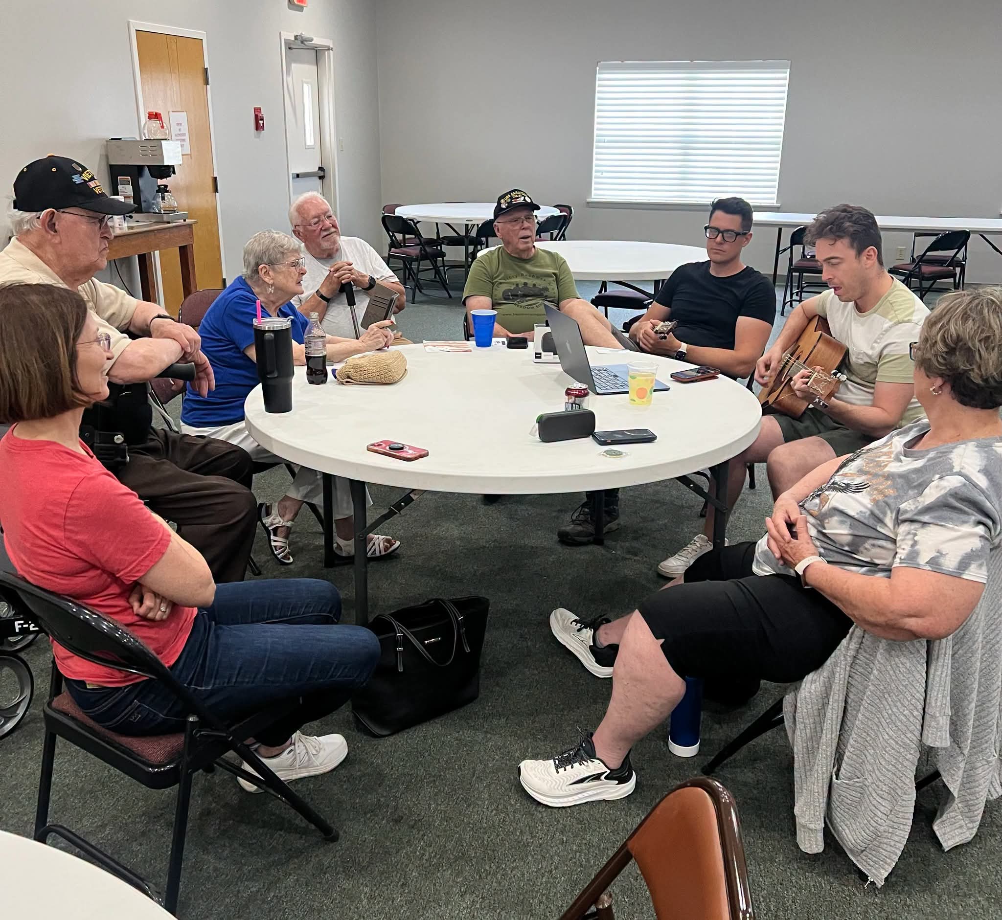 Group of people sitting around a round table in a room, engaging in conversation, with some holding cups.