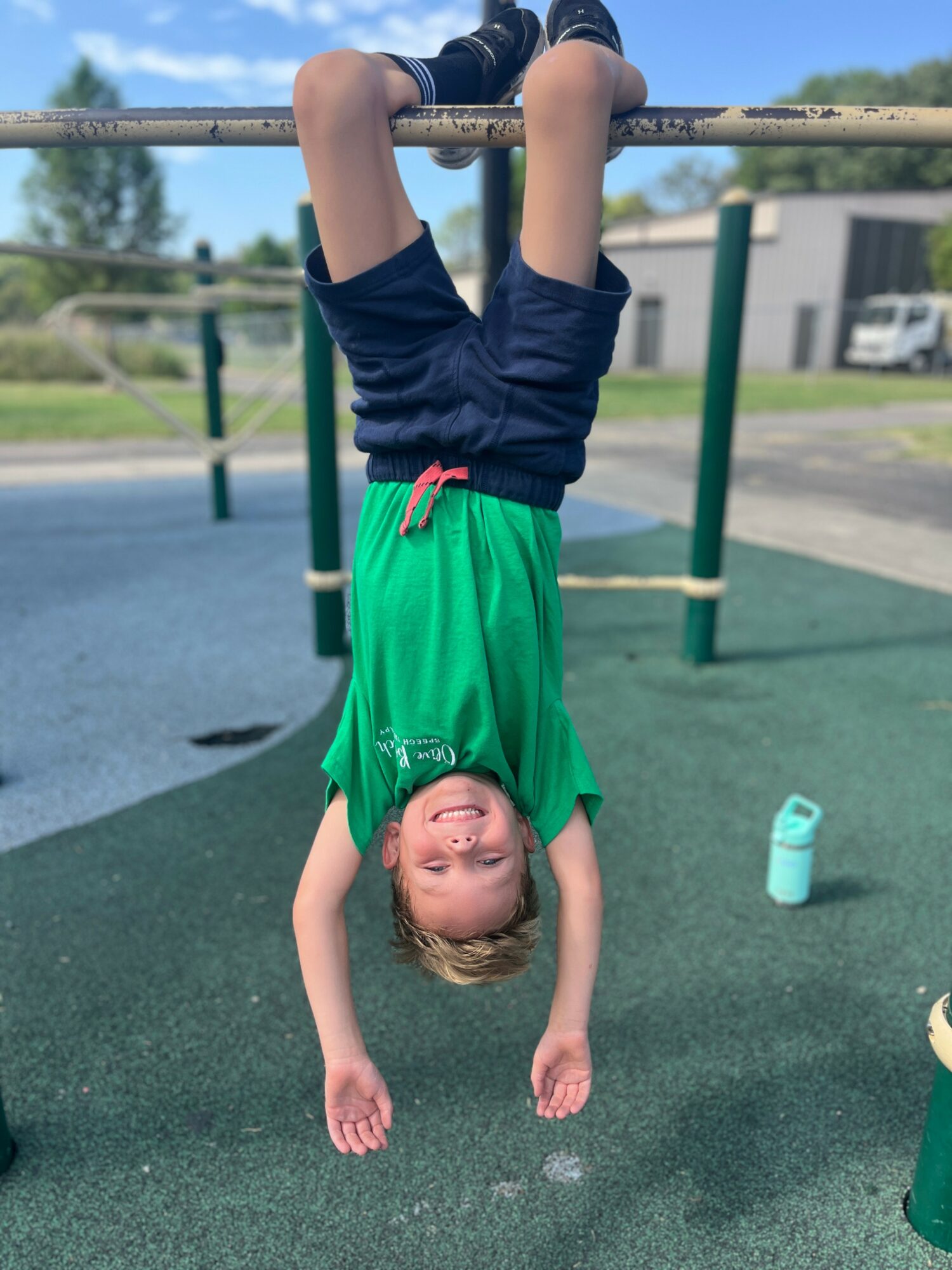 Child hanging upside down from a horizontal bar, smiling, with a water bottle nearby, outdoors on a playground.