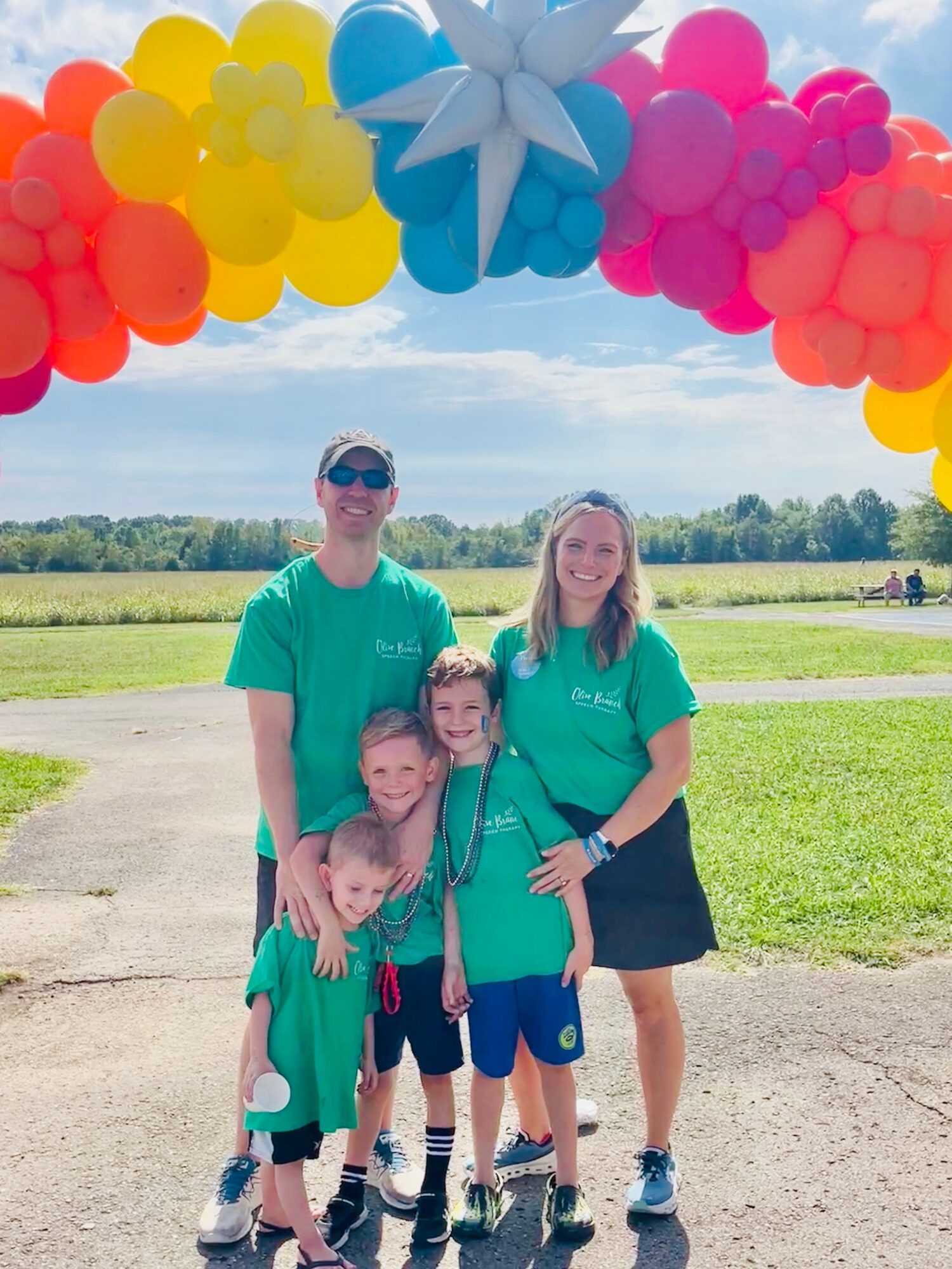 Four people standing outdoors under colorful balloon arch, smiling, with a grassy field and trees in the background.