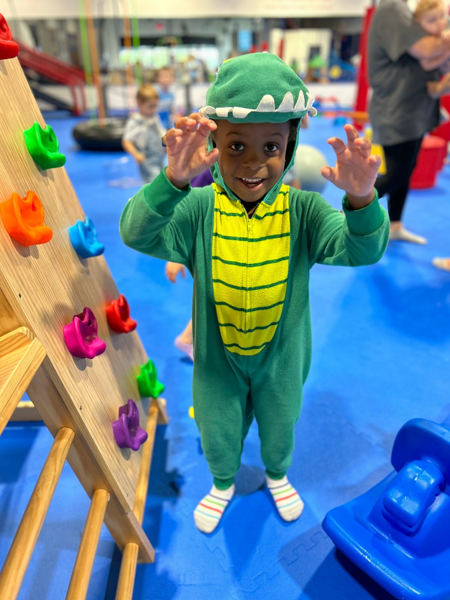 Child in a green dinosaur costume with a yellow striped belly, smiling and waving indoors on a blue floor.