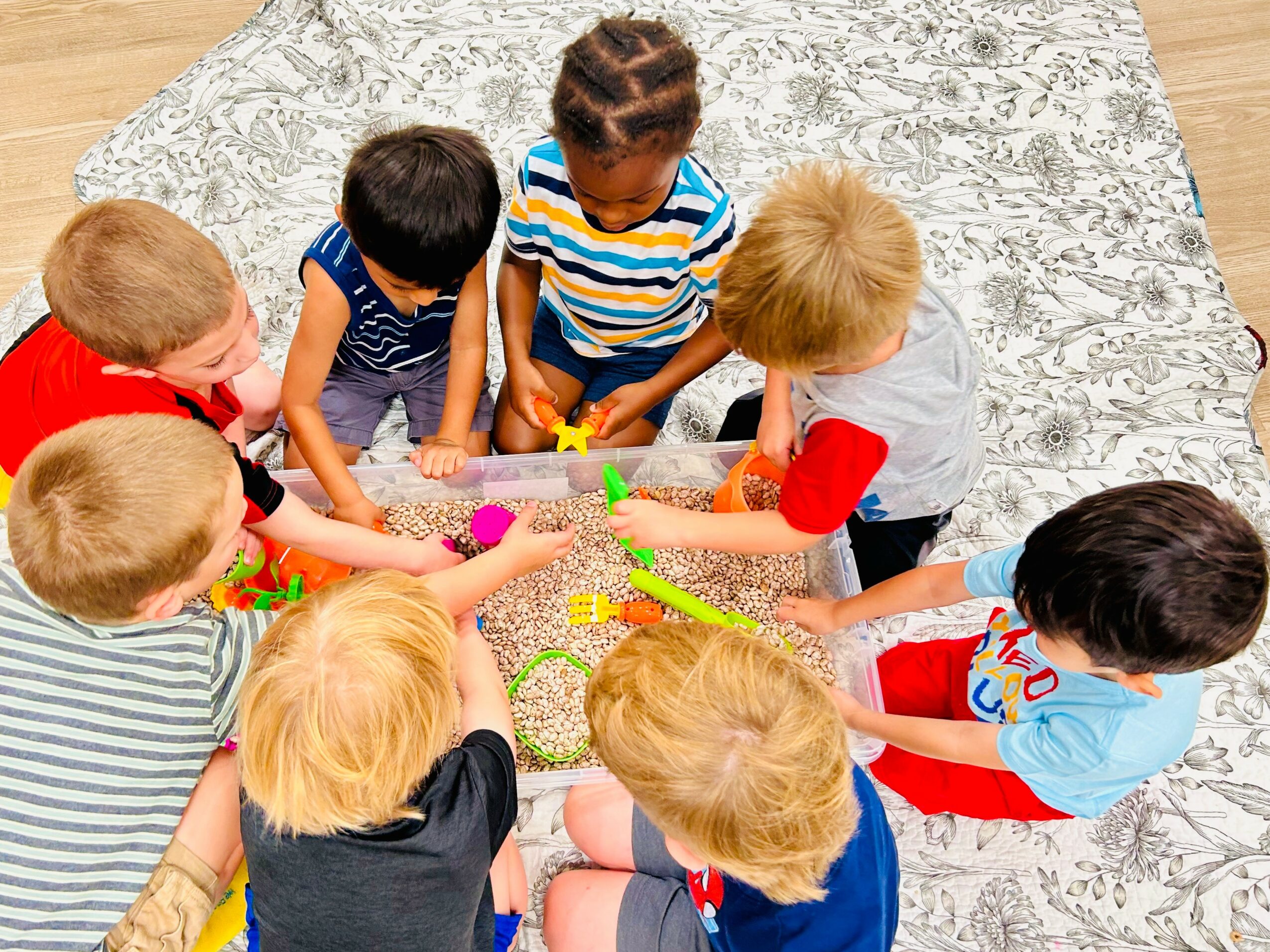 Children gathered around a game on a patterned rug, reaching toward colorful game pieces, viewed from above.