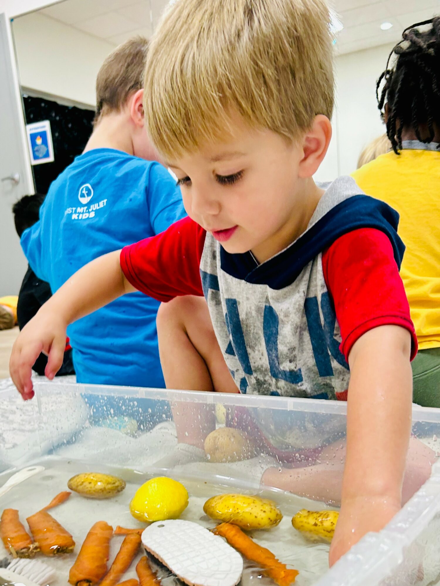 Young boy with blond hair reaches into a clear container with snacks, surrounded by children in a classroom.