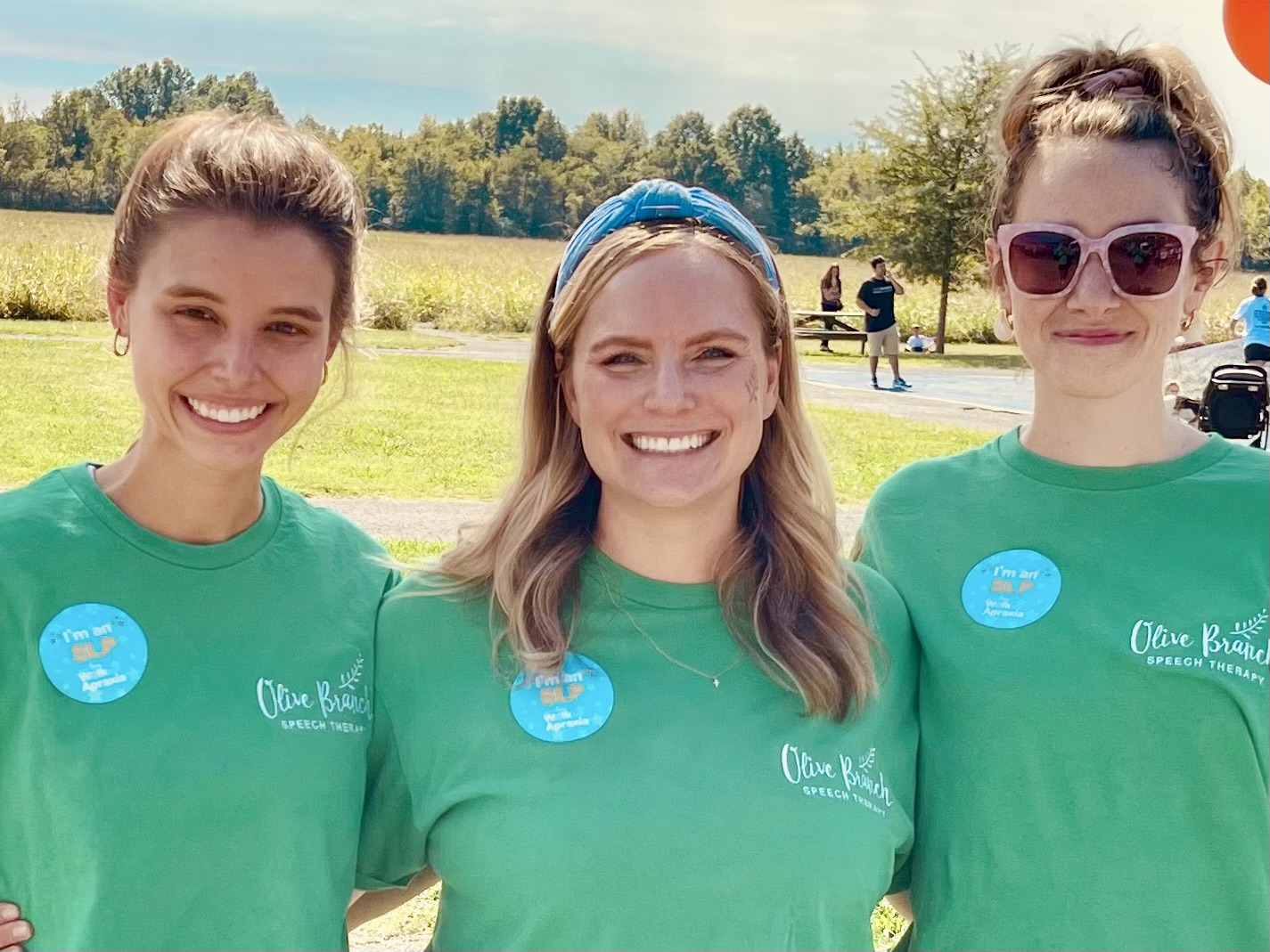 Three women smiling outdoors wearing matching green shirts with a blue badge, standing in a grassy area with trees and people in the background.