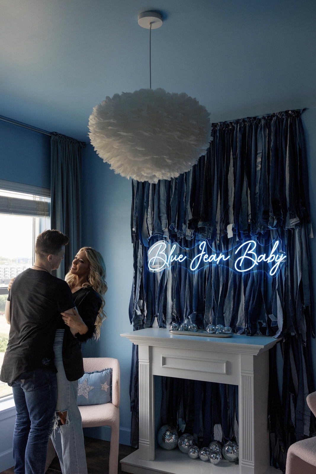 Two people standing near a window with blue curtains, a white fireplace, and a neon sign that reads 'Blue Jean Baby'.
