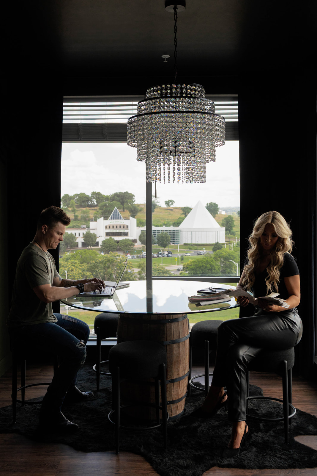 Two women sit at a round table near a large window with a chandelier overhead, overlooking a landscape with buildings and trees.