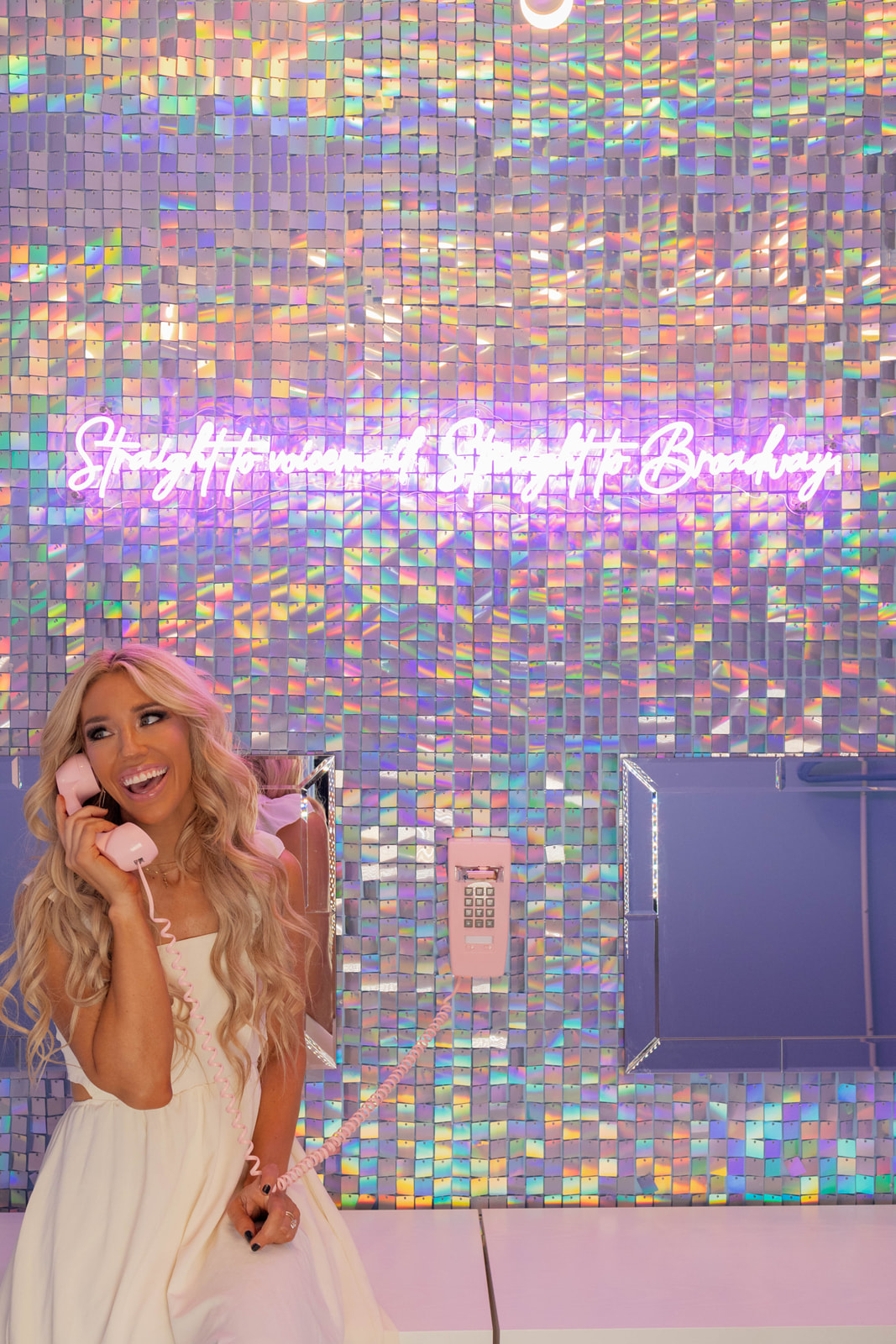 Woman with long blonde hair in a white dress smiling, background with colorful mosaic wall and neon sign.