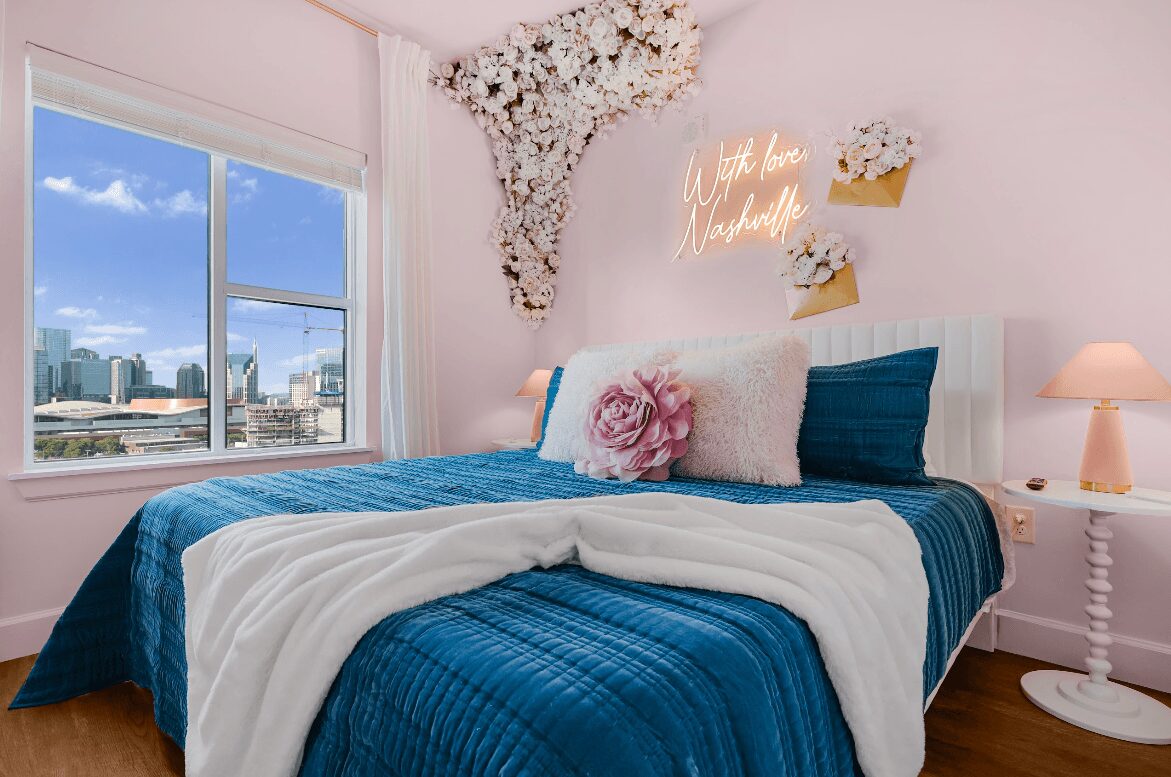 Bedroom with a large window, bed with blue bedding, pink wall, floral wall decor, and a pink lamp on a white side table.