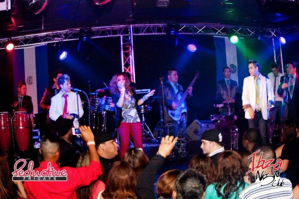 Band performs on stage with colorful lights, audience watching and taking photos, some raising hands, lively atmosphere.