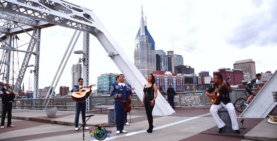 Group of people performing music on a bridge with city skyline in background, including a tall building with spire.