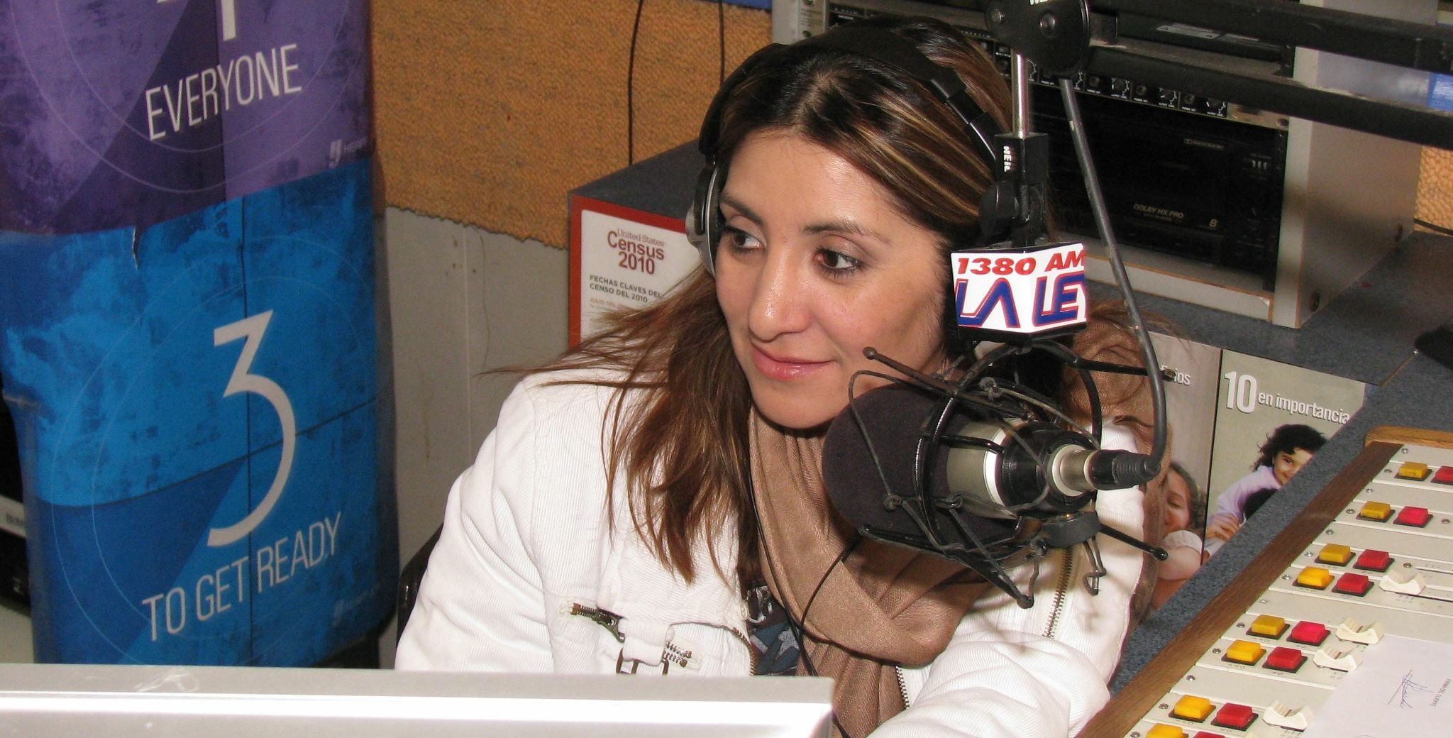 Woman with long hair wearing white jacket speaking into a microphone in a radio studio.