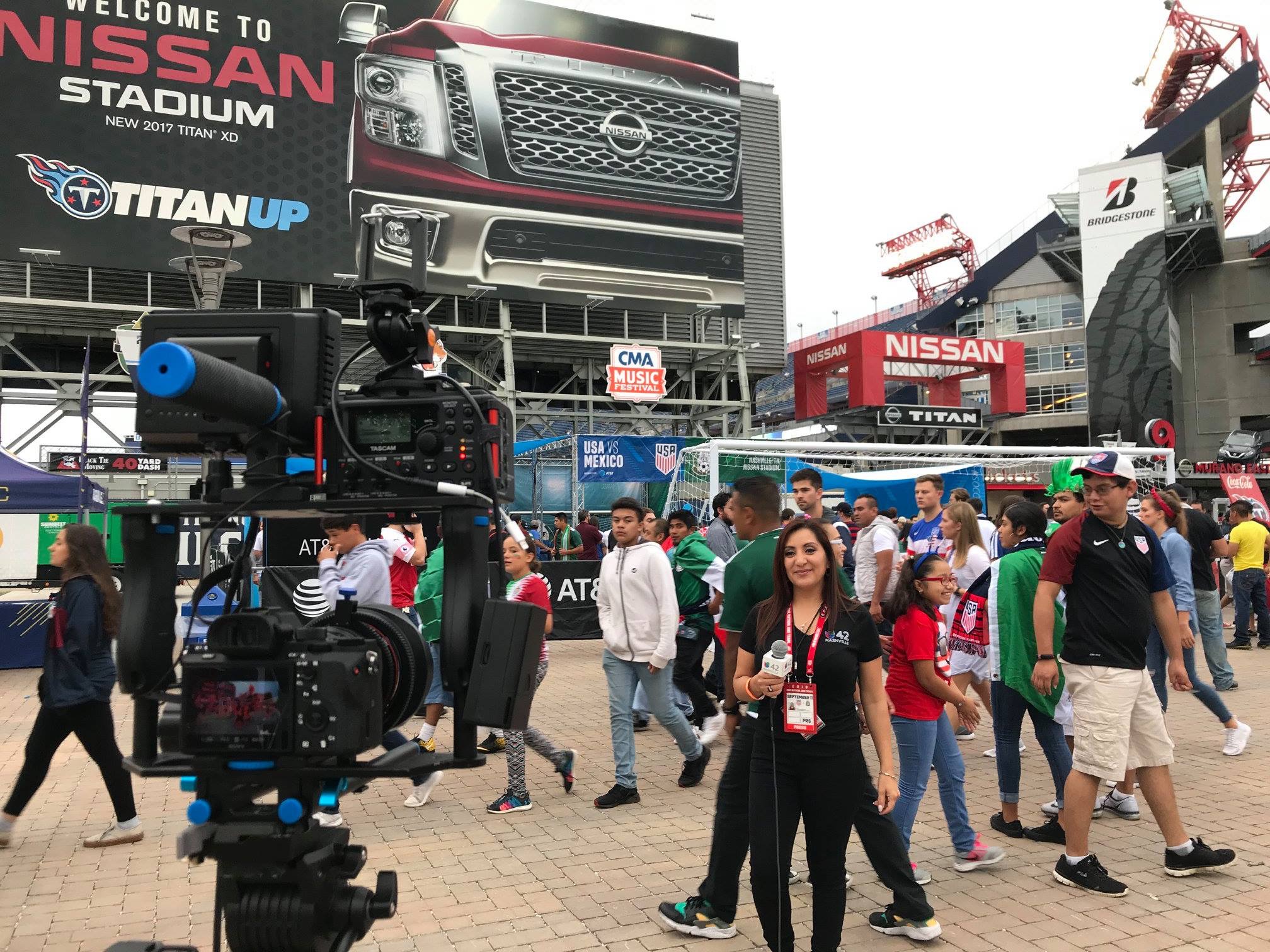 Crowd of people walking at an outdoor Nissan event with large Nissan banners and a camera setup in the foreground.