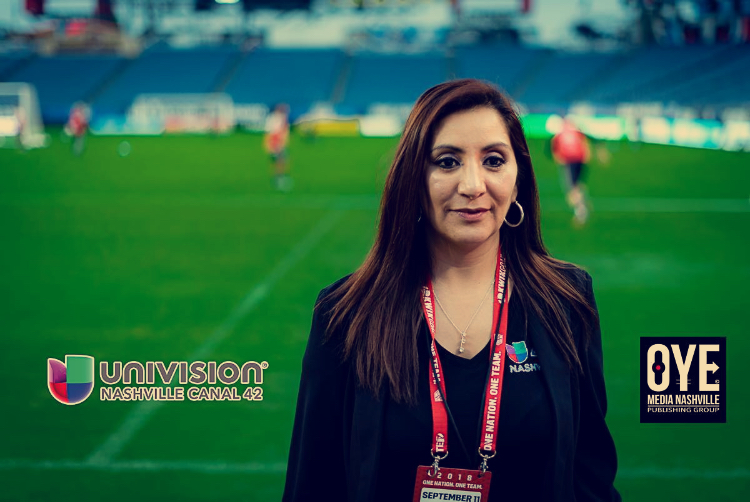 Woman with long dark hair and hoop earrings standing on a sports field, smiling, with a stadium background.