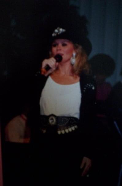 Woman singing into a microphone, wearing a hat, earrings, and a black and white outfit, in a dark setting.