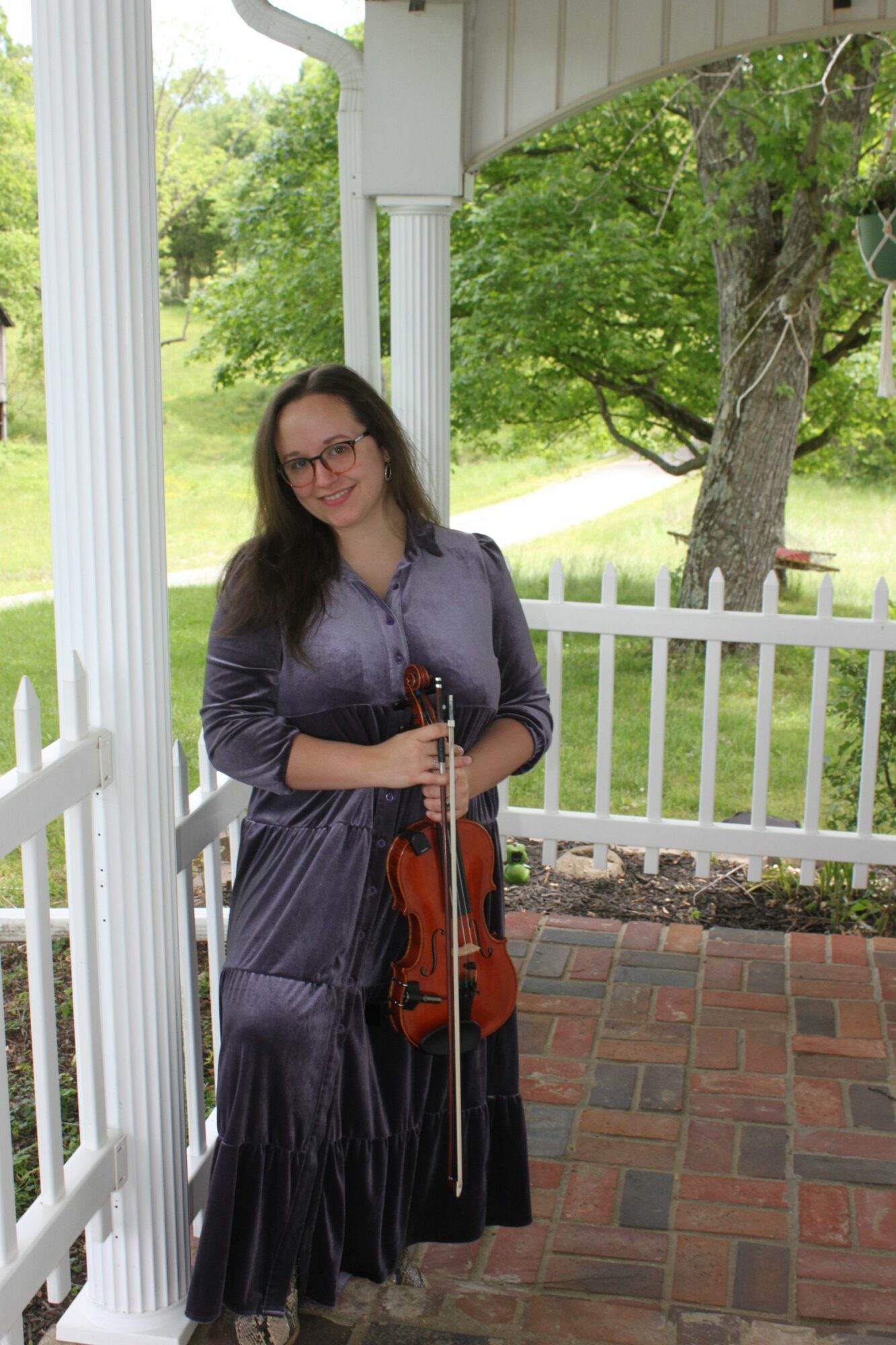 Woman holding a violin and bow, standing outdoors on a porch with white railing and green trees in background.