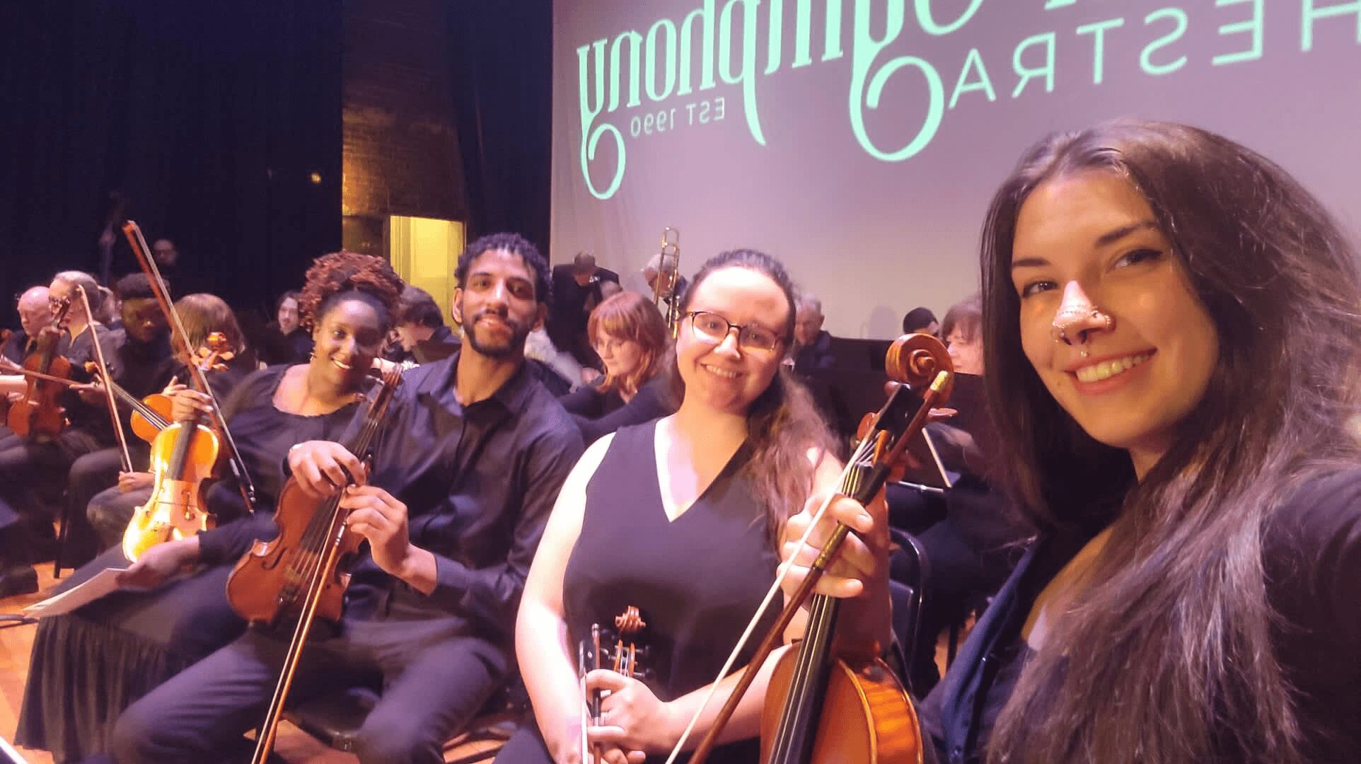 Group of musicians holding violins on stage, smiling, with a screen in the background displaying text.