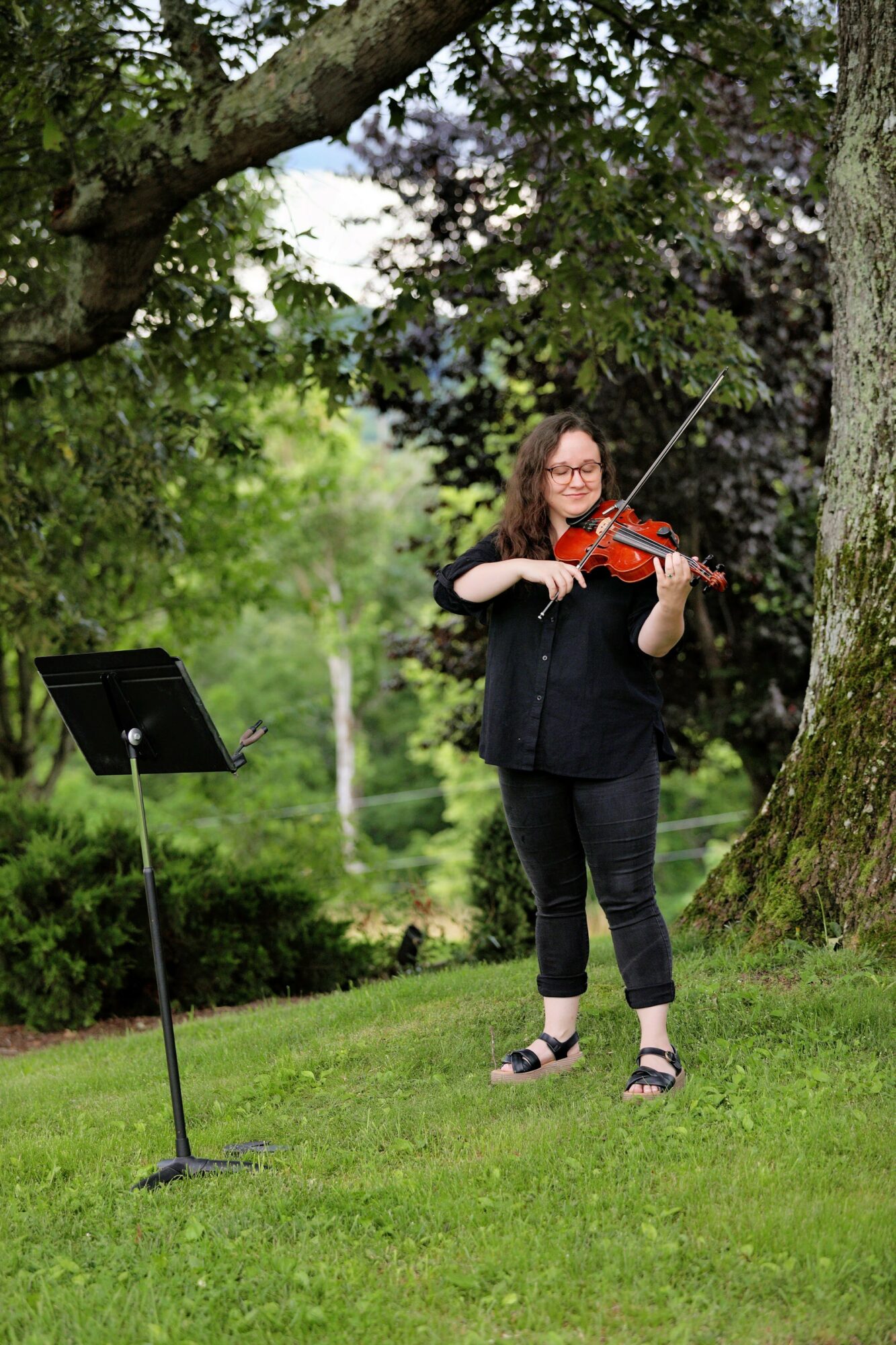 Woman playing violin outdoors under a tree, music stand nearby, green grass and trees in background.