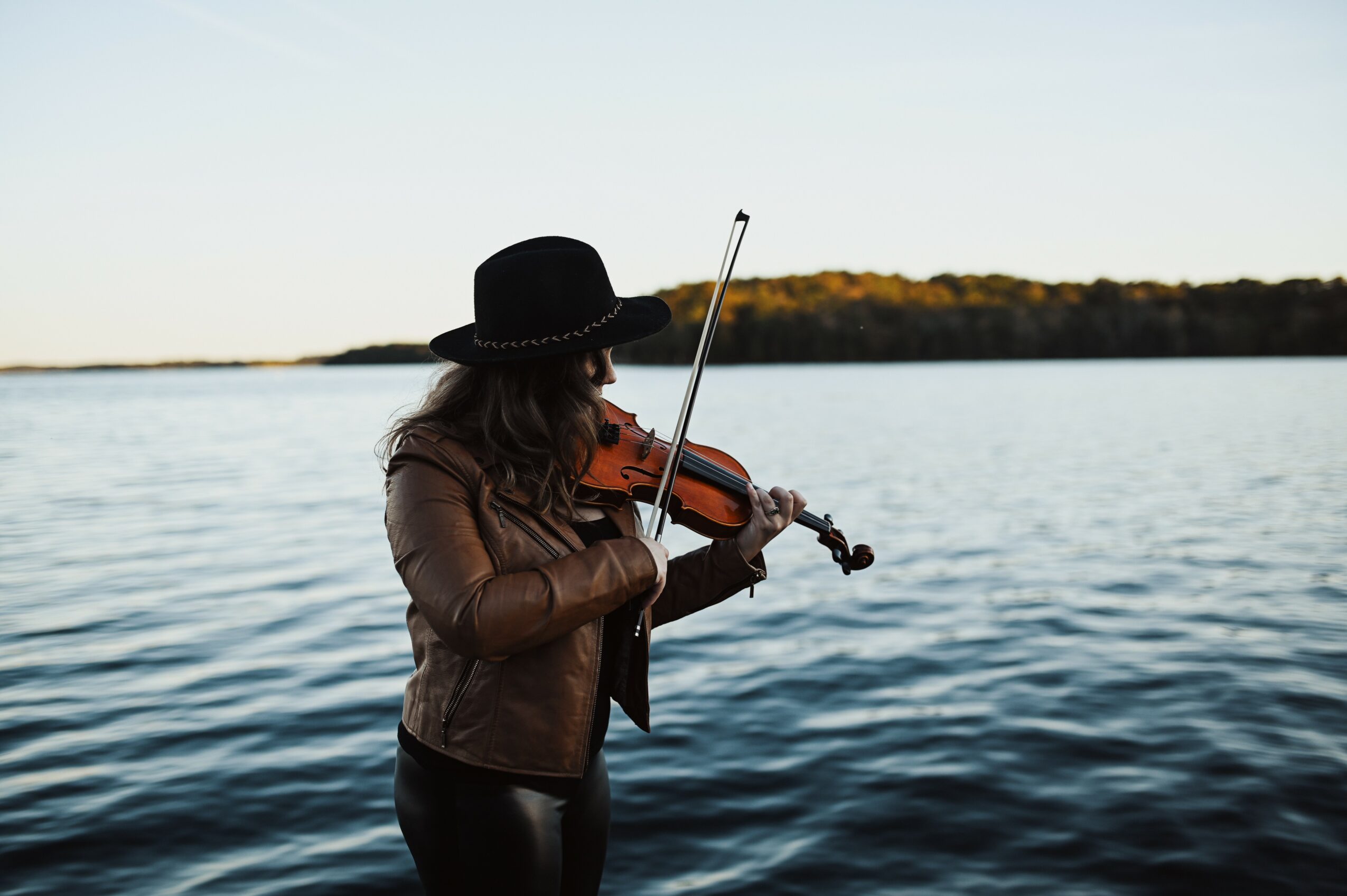 Person wearing a hat playing a violin by a body of water with a distant shoreline, during daytime.