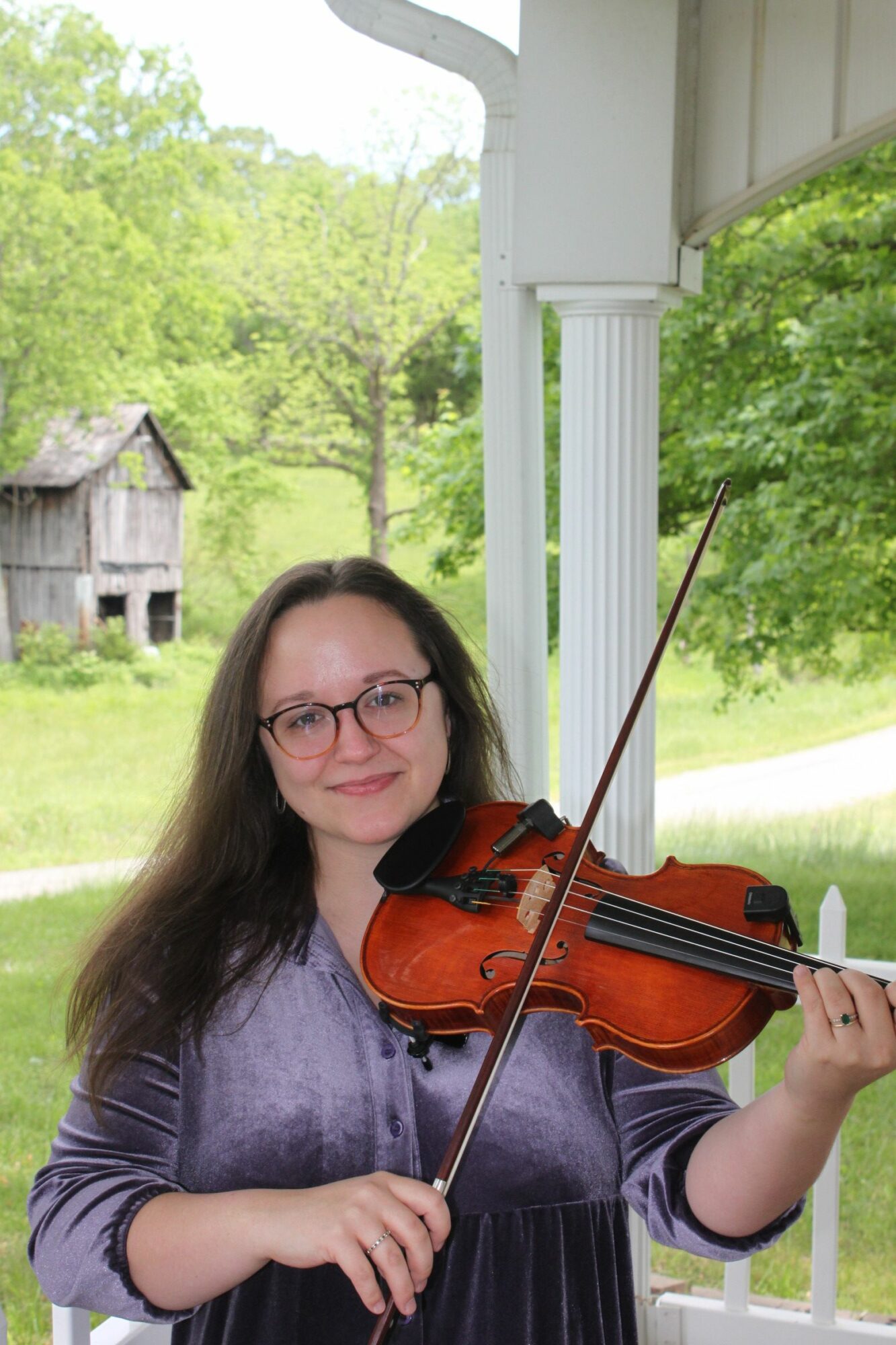 Woman with glasses playing violin outdoors, smiling, with green trees and a white porch in the background.