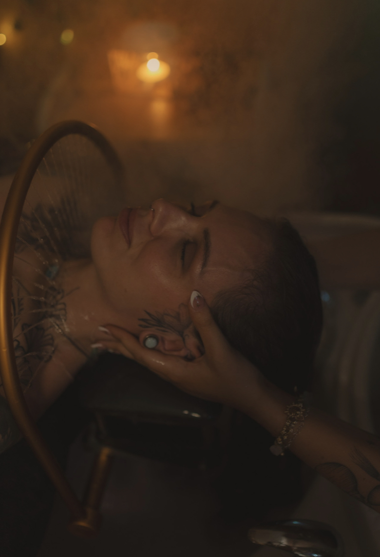 Person with wet hair relaxing in a dimly lit setting, with a candle in the background, eyes closed, hand on face.