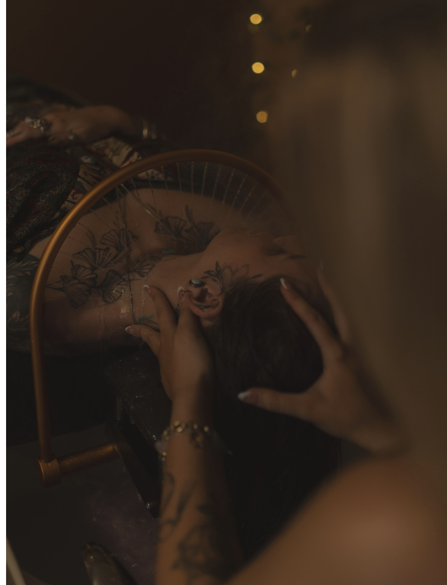 Person lying down with hands on head, tattoos visible, in a dimly lit environment with warm lights in the background.