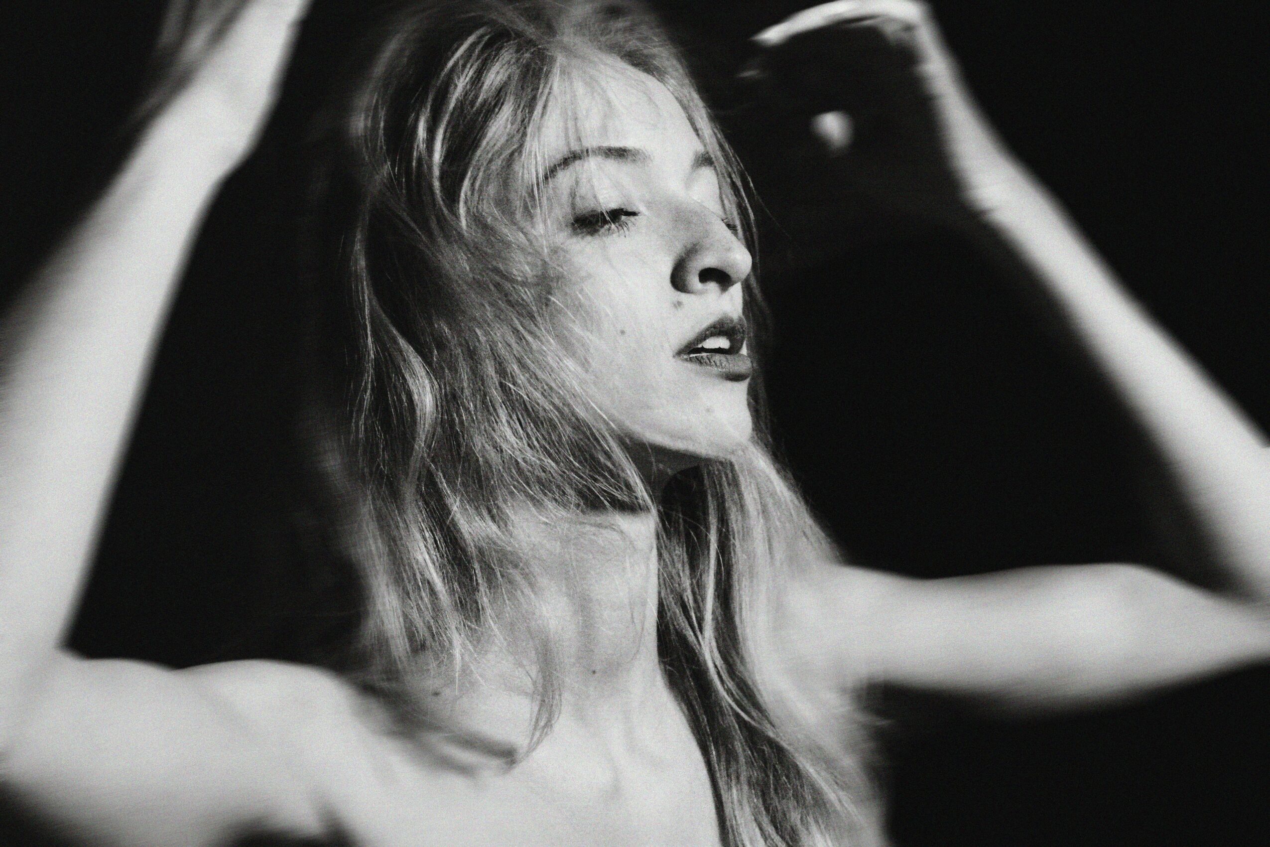 Woman with long hair, eyes closed, head tilted, arms raised, in black and white.