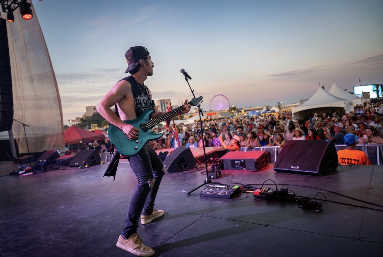 Female musician playing guitar on stage at outdoor concert during sunset, with crowd and tents in background.