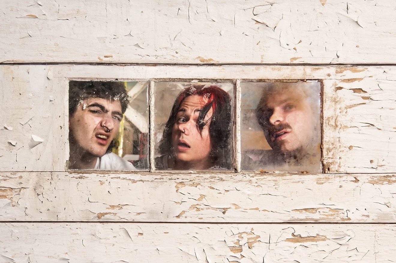 Three people look through a small horizontal window in a weathered white wooden wall.