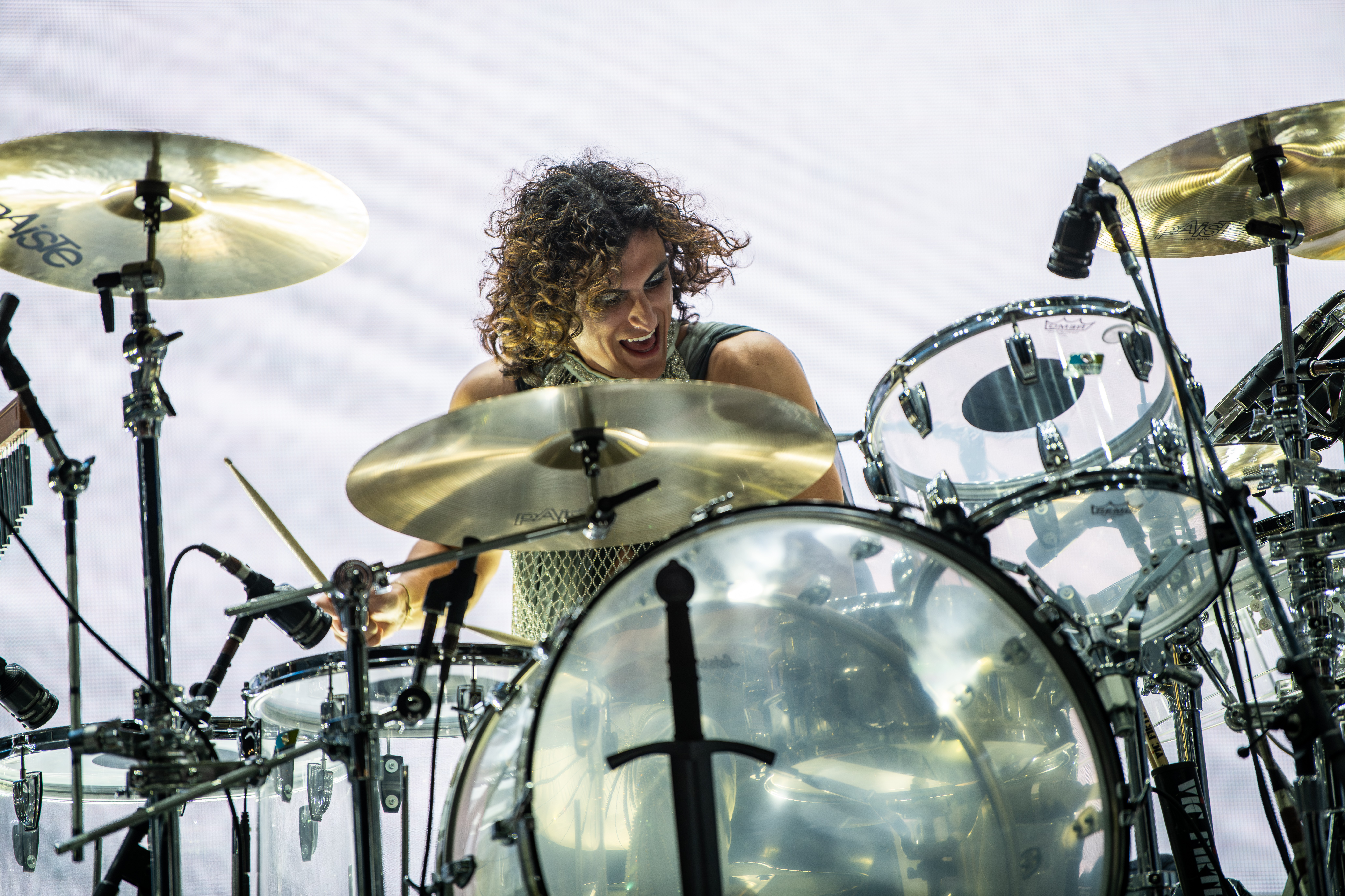 A woman with curly hair playing drums on stage, surrounded by cymbals and drum equipment, smiling.