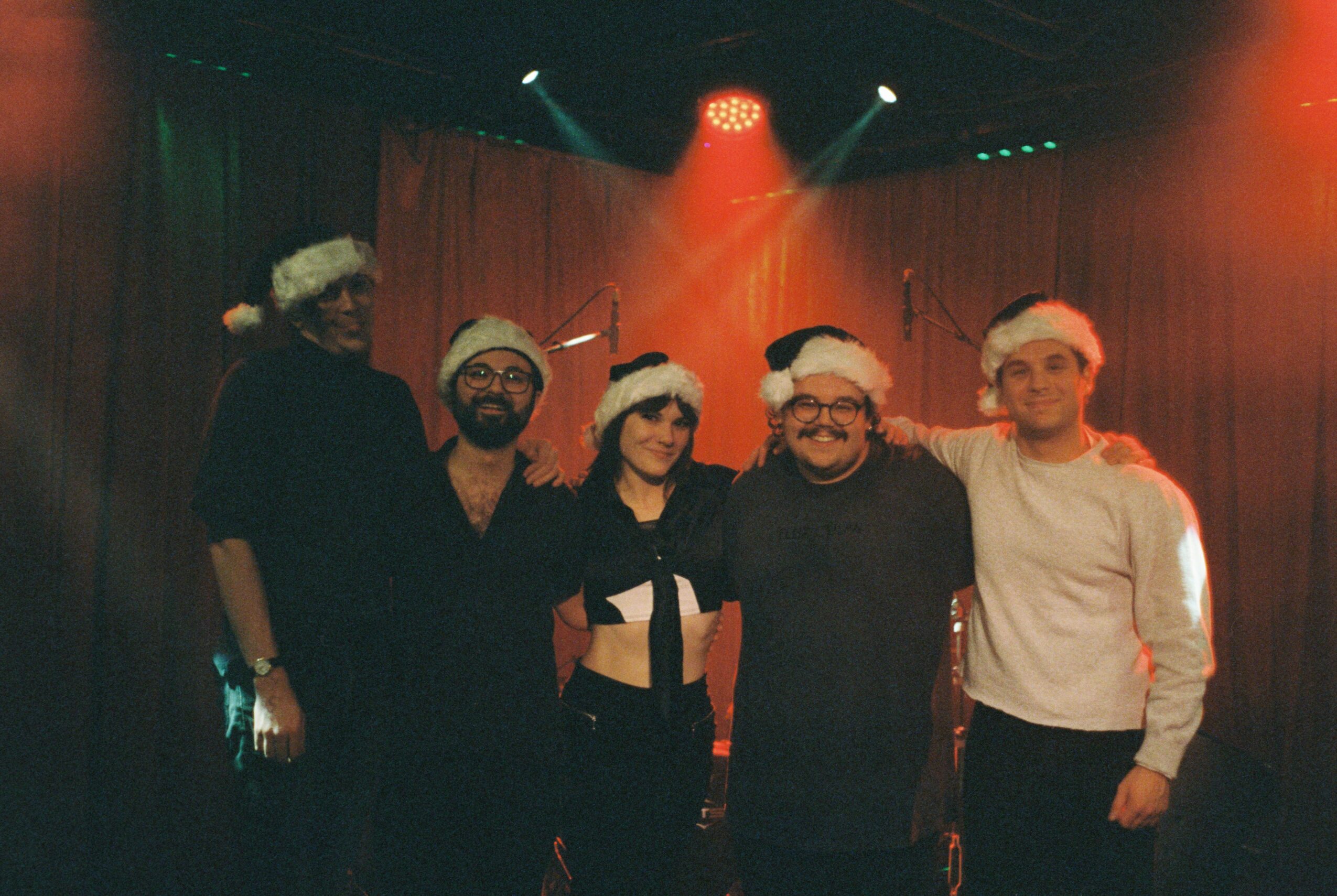 Five people wearing Santa hats standing on a stage with red curtains and stage lights.