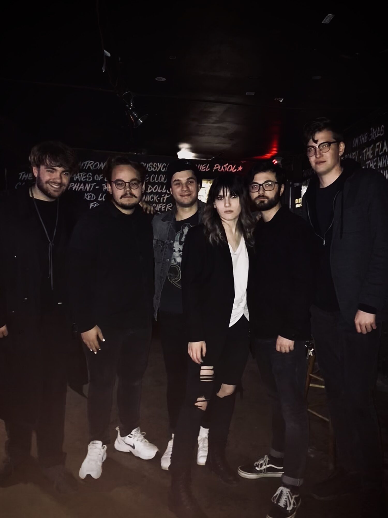 Group of six people standing together in a dark indoor setting, some wearing glasses and dark clothing.