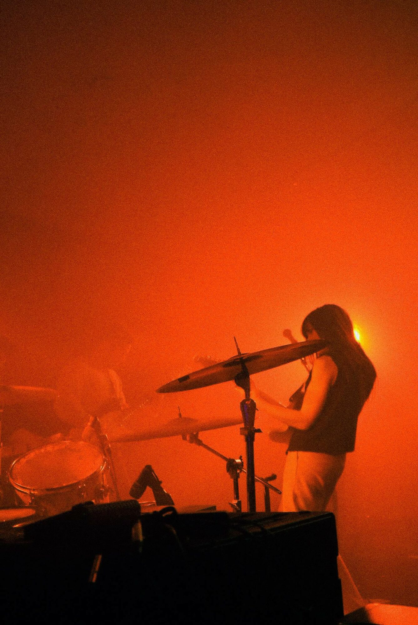 Silhouette of a person playing drums on stage with orange lighting and fog, drum set visible in foreground.