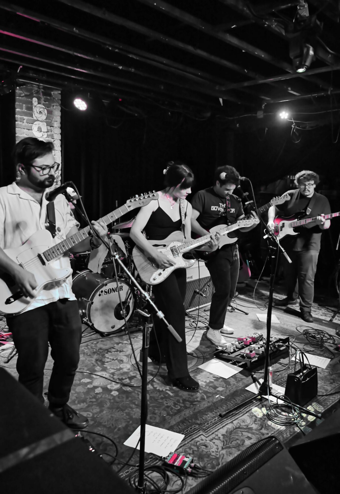 Five musicians perform on stage with guitars and drums, under stage lighting, in a dark venue with a brick wall backdrop.
