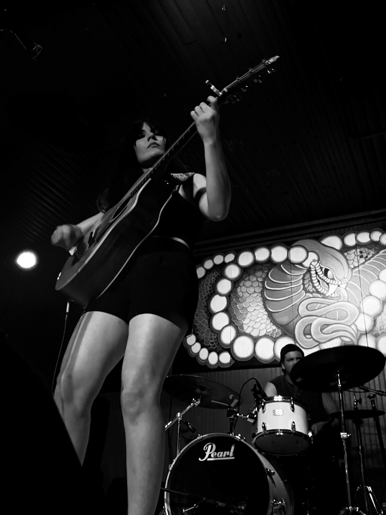 Female musician playing guitar on stage with a drum set and decorative backdrop, black and white image.