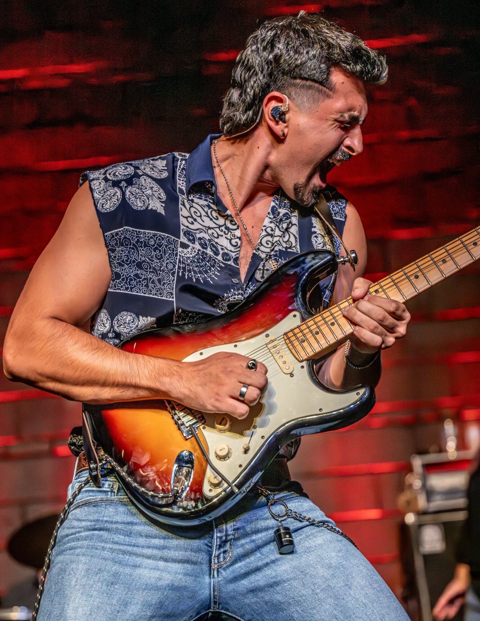 Person playing electric guitar passionately, wearing sleeveless shirt and jeans, with a red background.