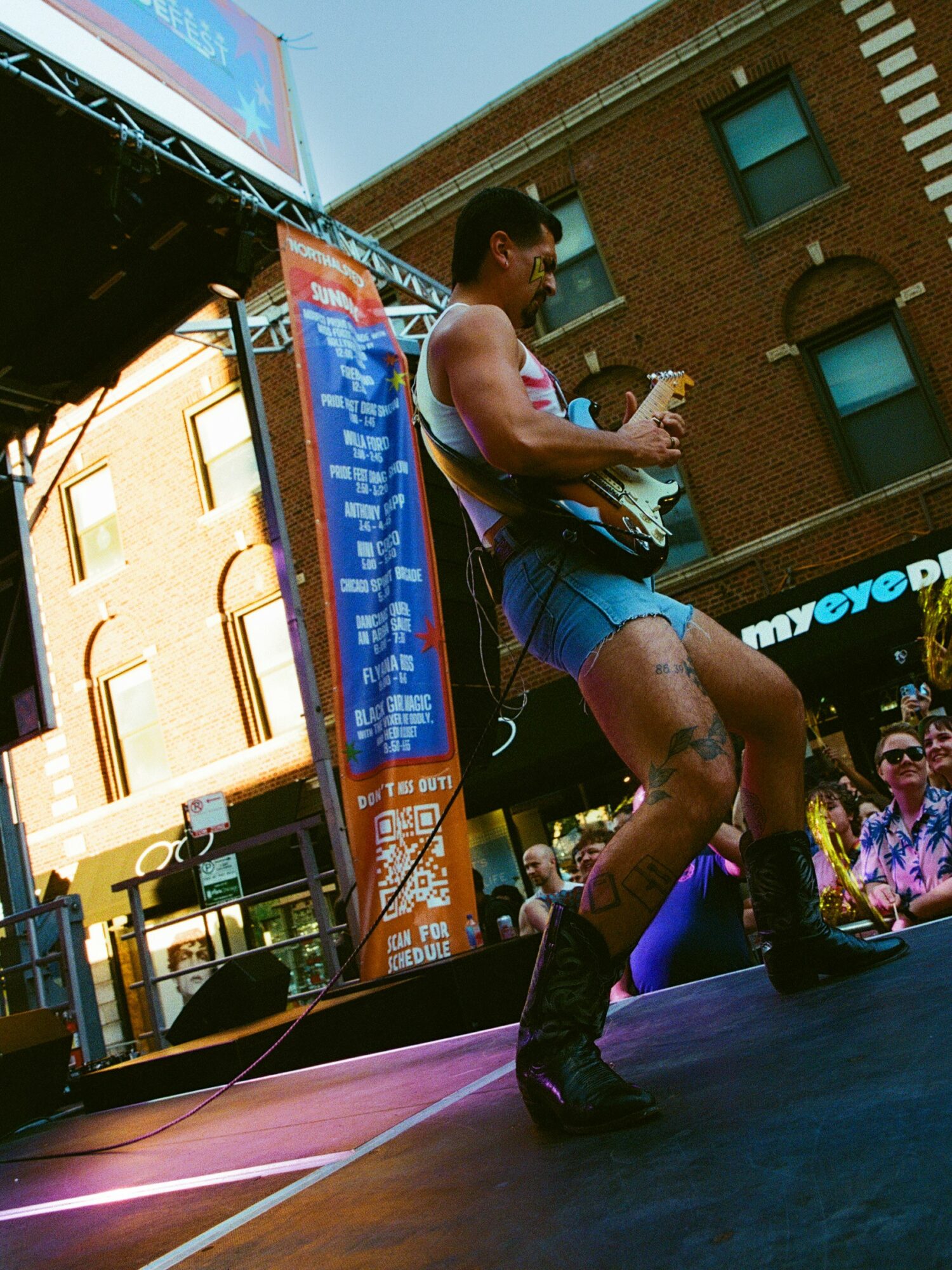Person dancing on stage with a guitar, wearing shorts and boots, in an outdoor urban setting with buildings and spectators.