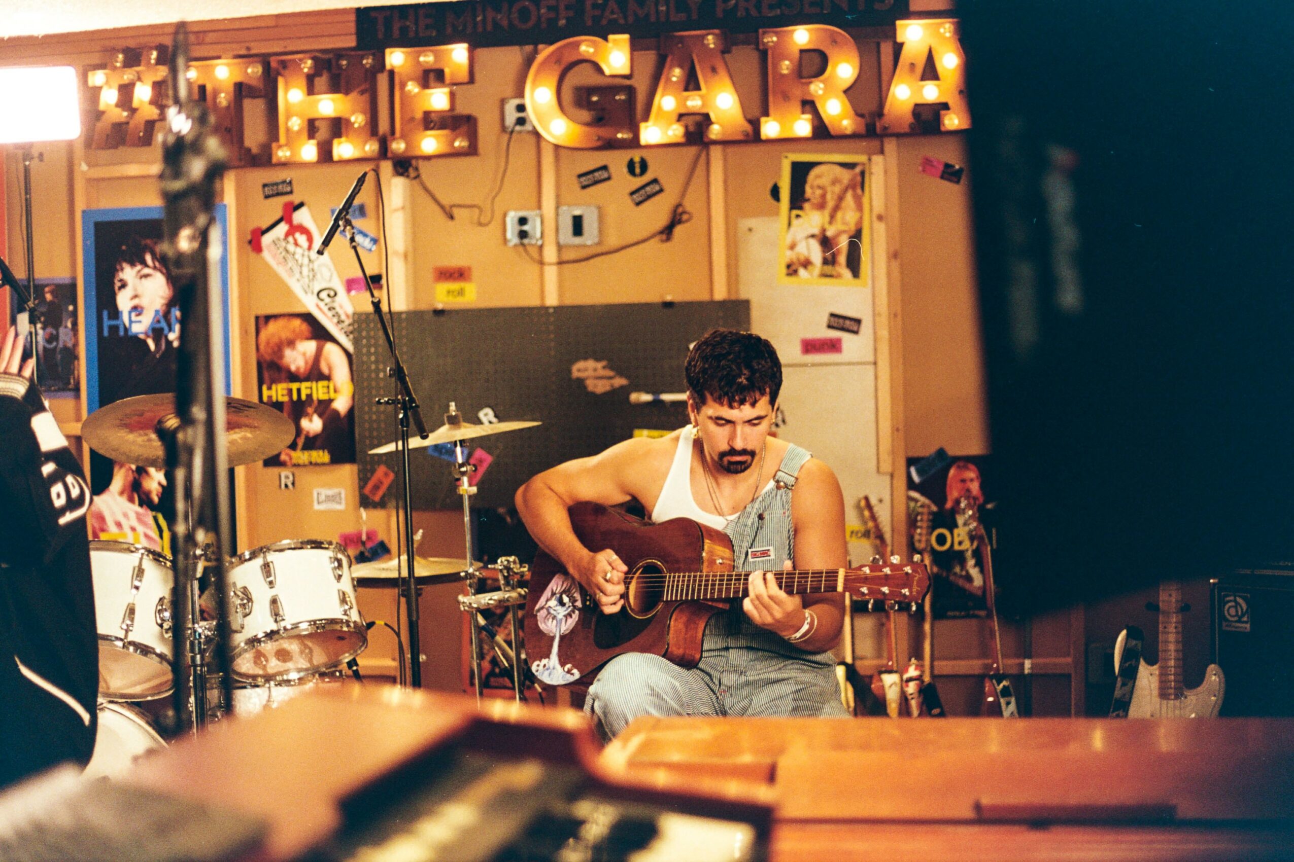 Person playing guitar on stage with drums and colorful posters in background, illuminated sign reading 'THE GARA'.