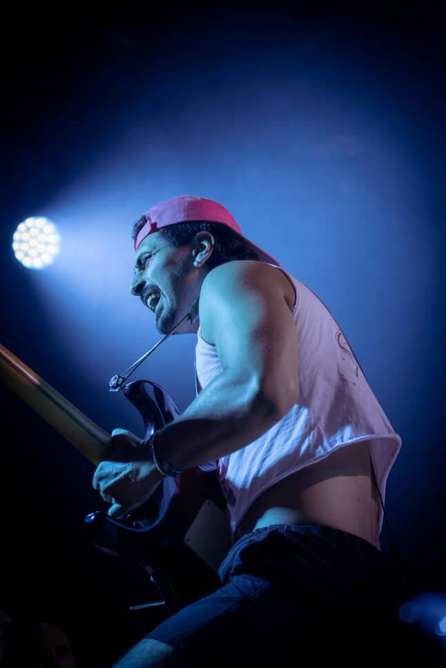 Person playing guitar on stage with blue lighting, wearing a pink cap and sleeveless shirt.
