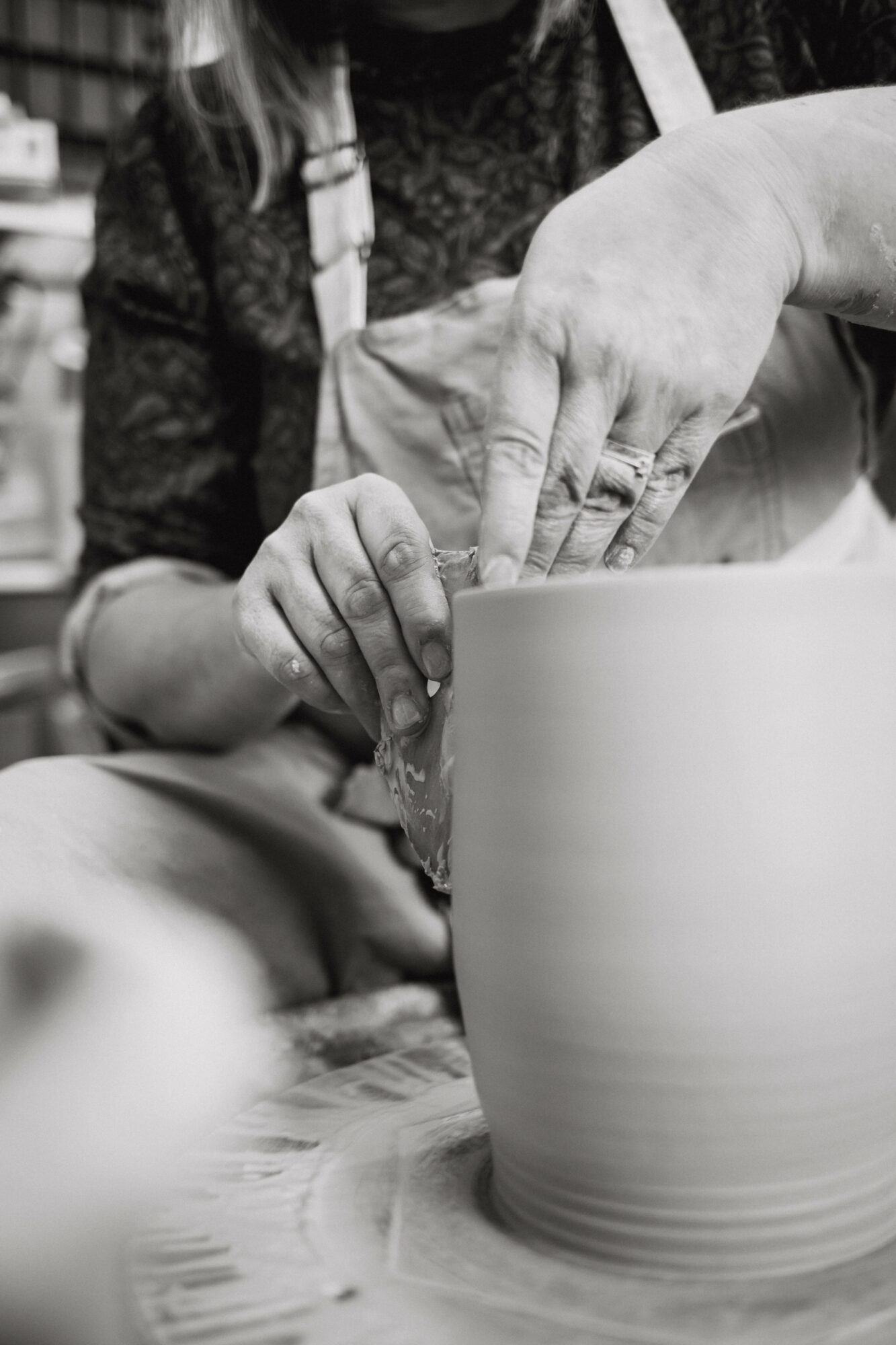 Two people work together on a pottery wheel, shaping a large clay vessel with their hands.