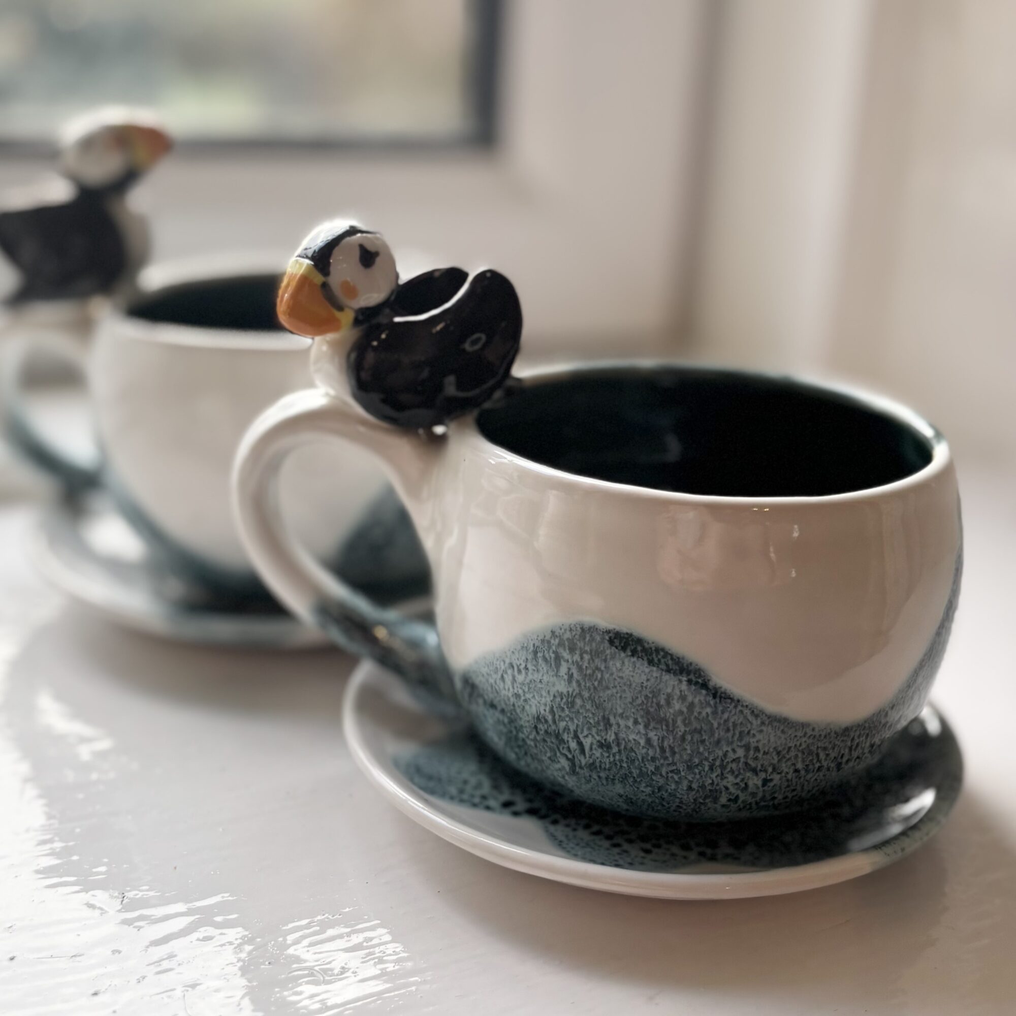 Two ceramic cups with bird-shaped handles on saucers, placed on a windowsill, with a blurred window in the background.