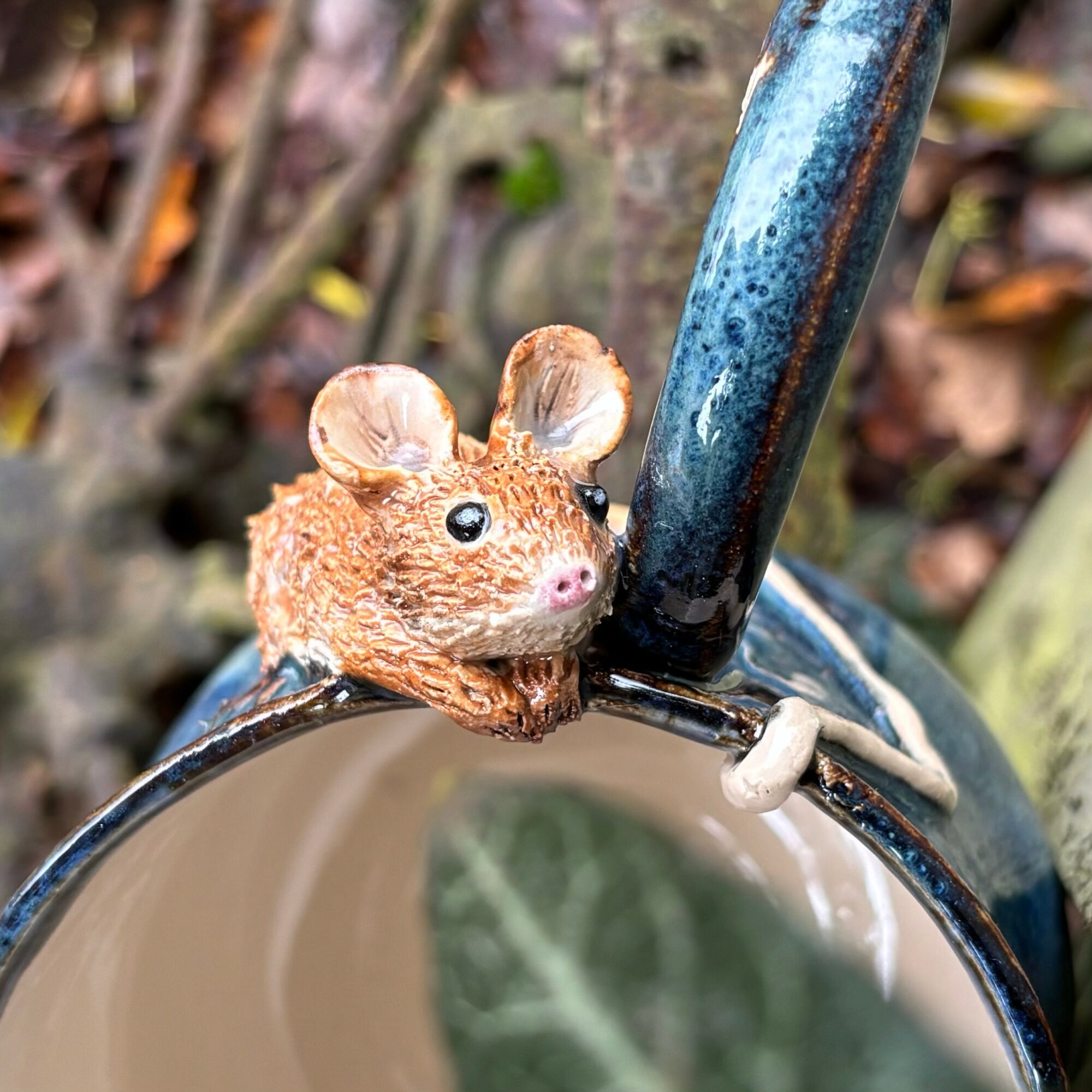 A small decorative mouse figurine with big eyes and pink nose, perched on a metal object outdoors.