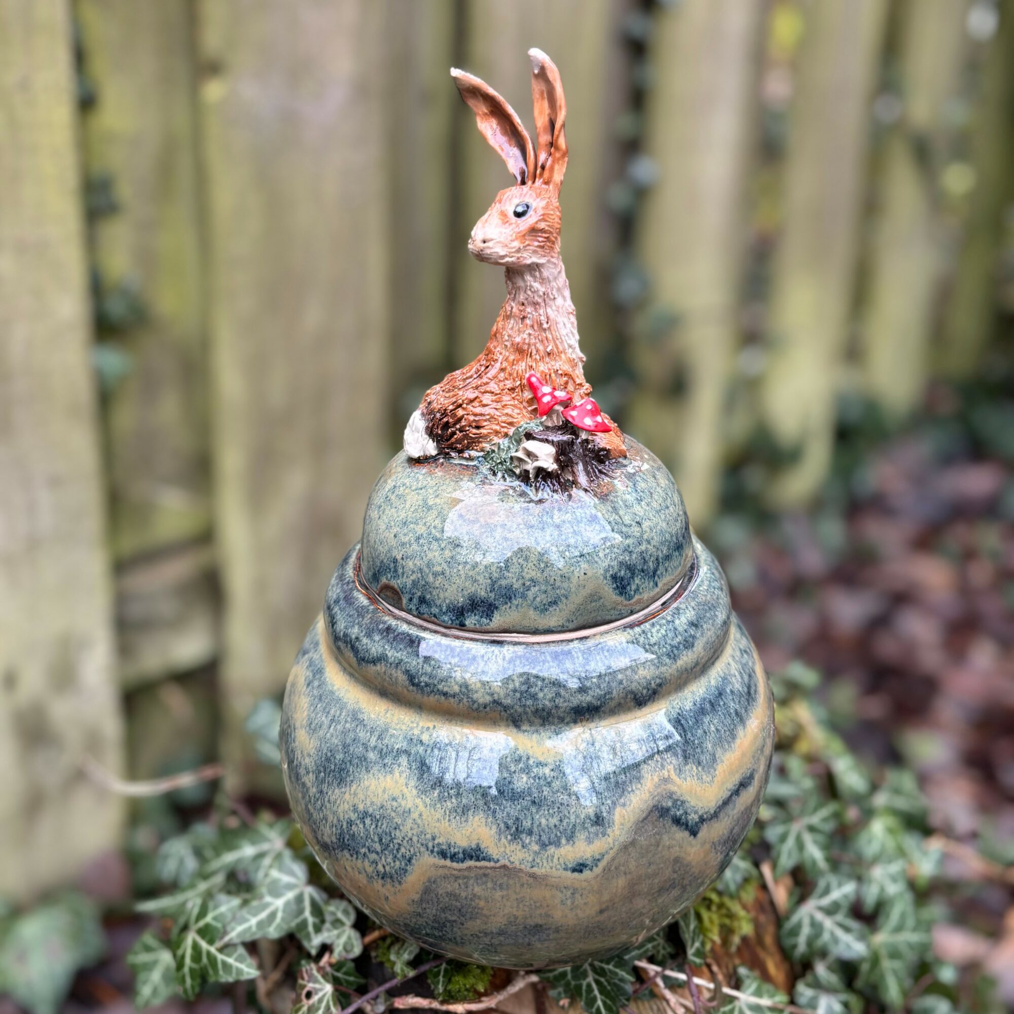 Decorative rabbit figurine on a rounded stone with plants and a wooden fence in background.
