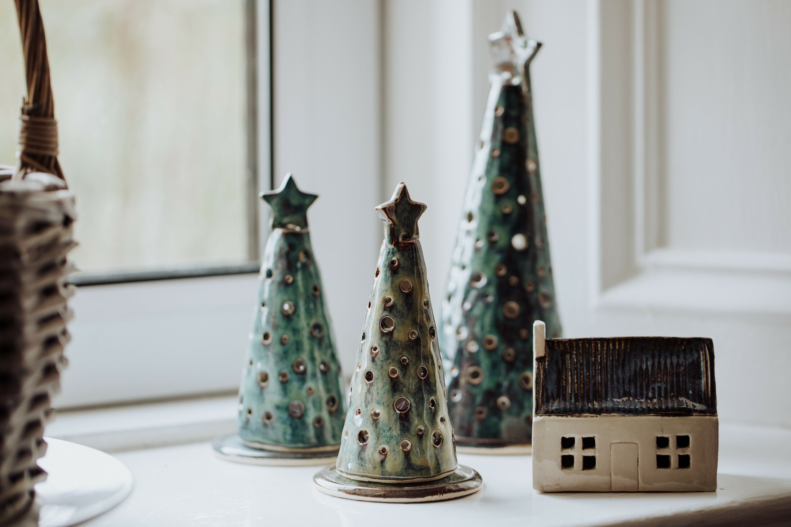 Three decorative Christmas trees and a small house figurine on a windowsill, with a window and wall in the background.