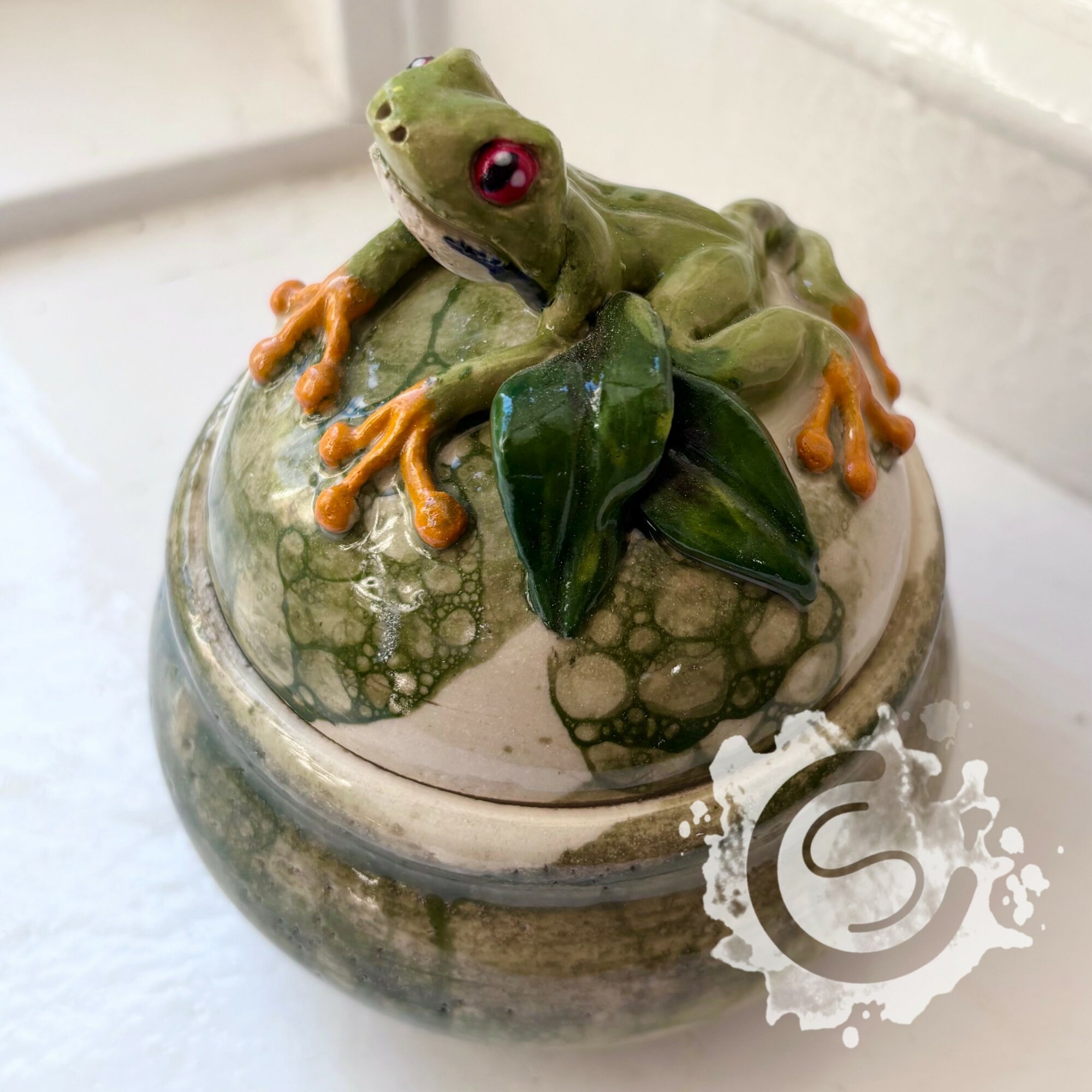 Decorative frog figurine with red eyes on a ceramic container with green leaves, textured surface, and a white background.