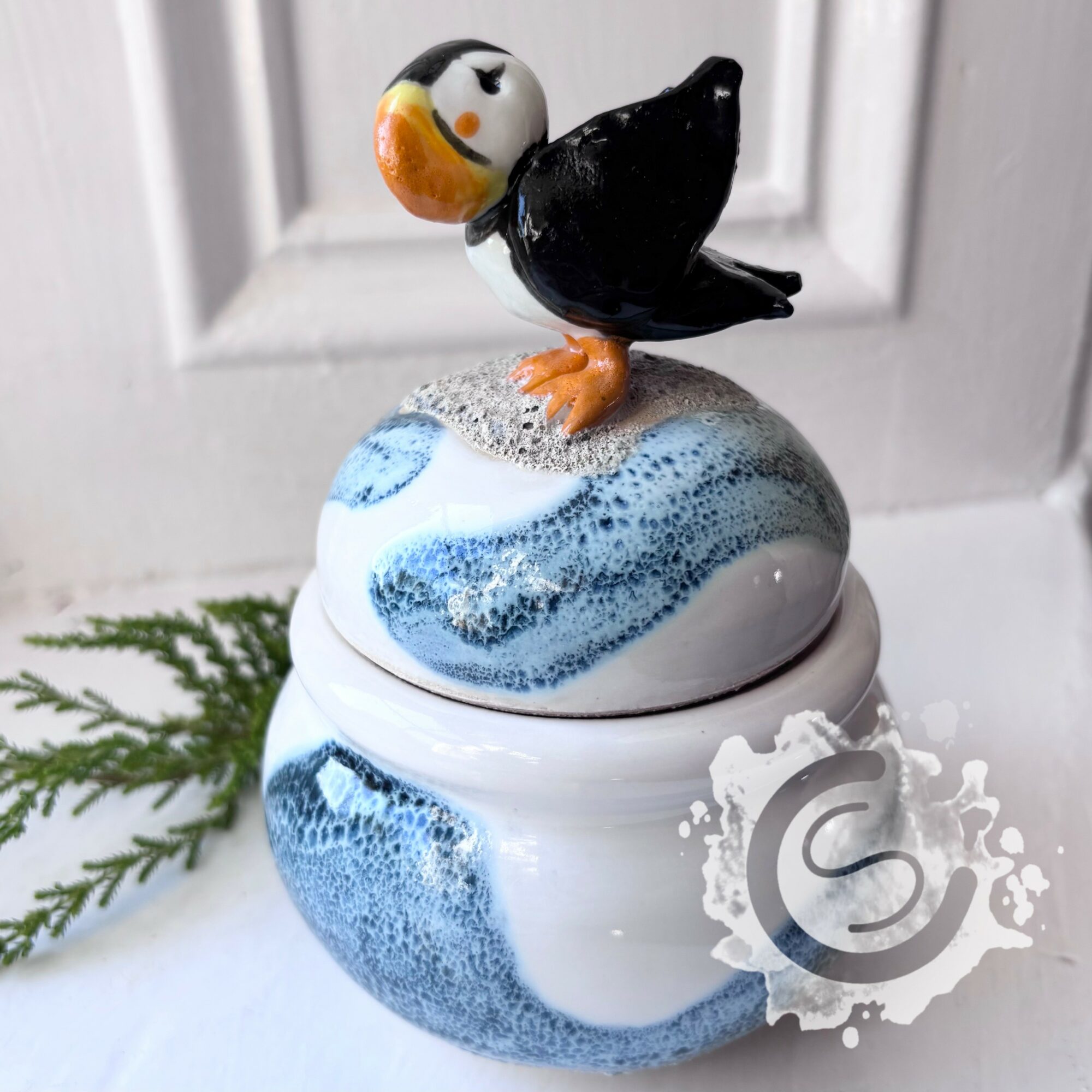 Decorative jar with a puffin figurine on top, featuring blue and white patterns, placed in front of a white door with greenery.